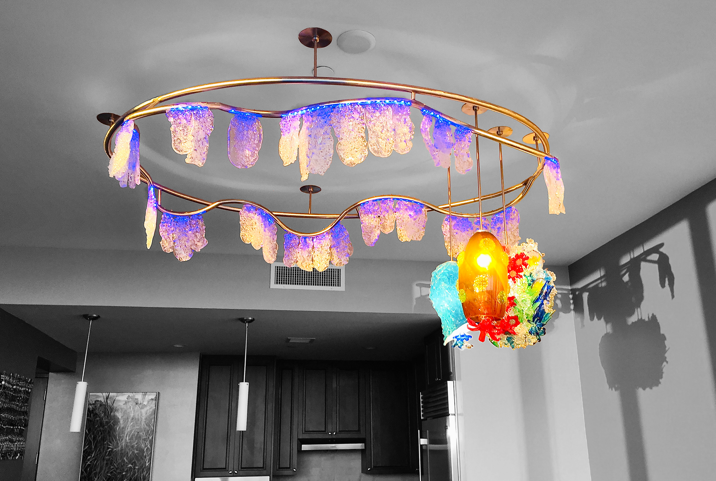 Colorful glass and LED chandelier with purple, blue, green, yellow, and red pieces, hanging above a kitchen with dark cabinets.