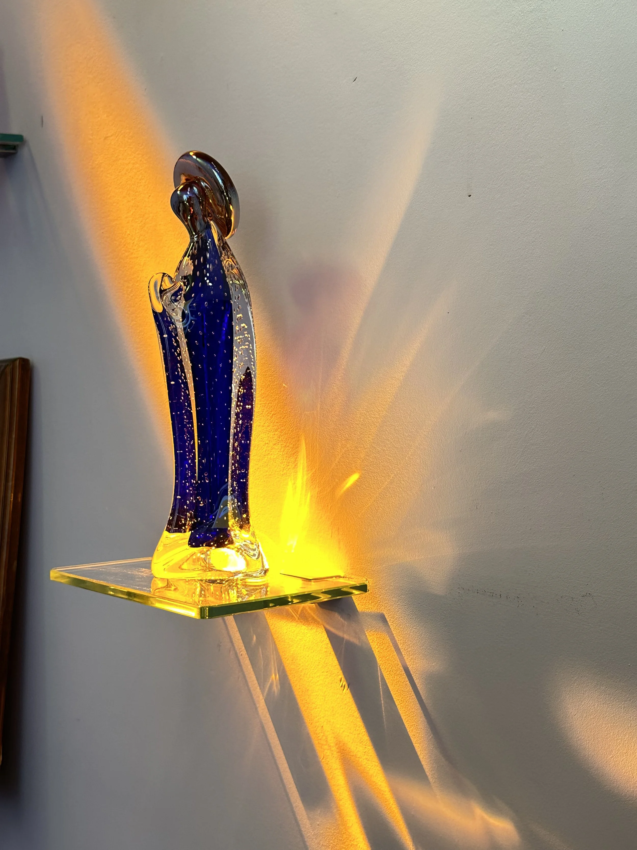 An abstract glass sculpture with blue and clear glass, illuminated by yellow lighting, casting colorful shadows and reflections on the wall.