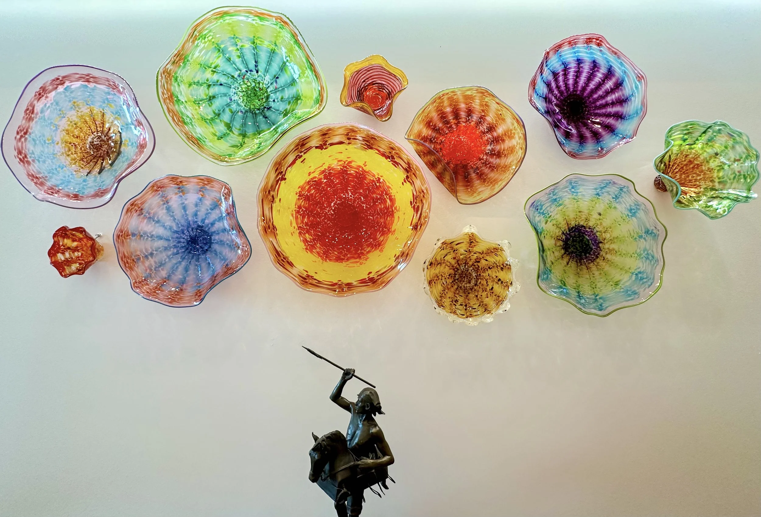 A modern kitchen with white cabinets, black countertops, and a backsplash. Two colorful, striped glass pendant lights hang from the ceiling. There are decorative plates and bowls on the counter and window ledge, with a window showing brickwork outside.