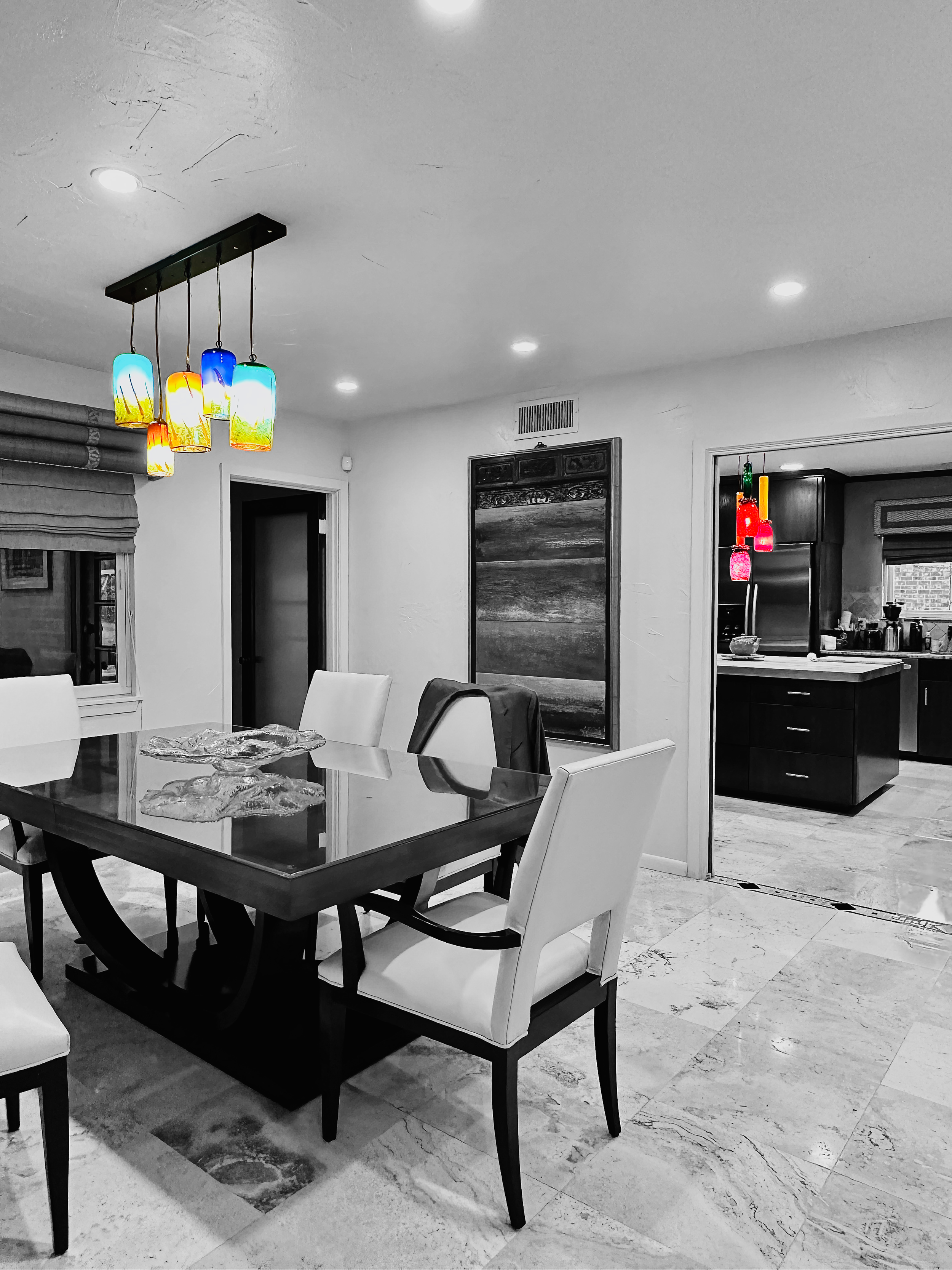 A dining room with a glass-top table surrounded by white cushioned chairs, colorful pendant lights hanging above, and a doorway leading to a kitchen. The room is primarily in black and white with the lights in color.
