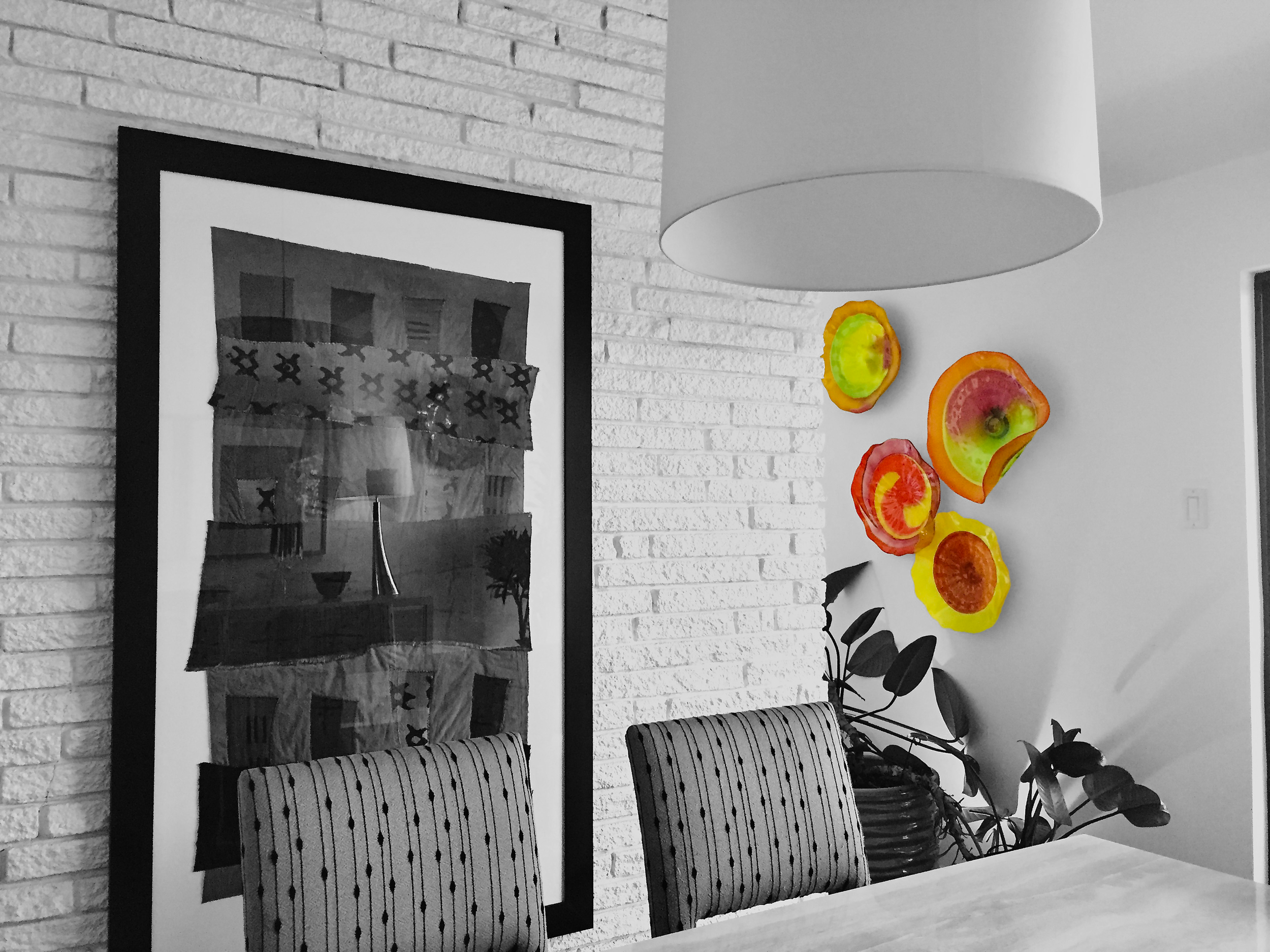 The image shows a dining area with black and white decor accented by colorful wall art. There are two striped chairs in front of a white table, a brick wall with a black frame picture, and vibrant, abstract, circular wall sculptures in yellow, orange, red, and green. A large white pendant lamp hangs above the table, and a potted plant is seen in the background.