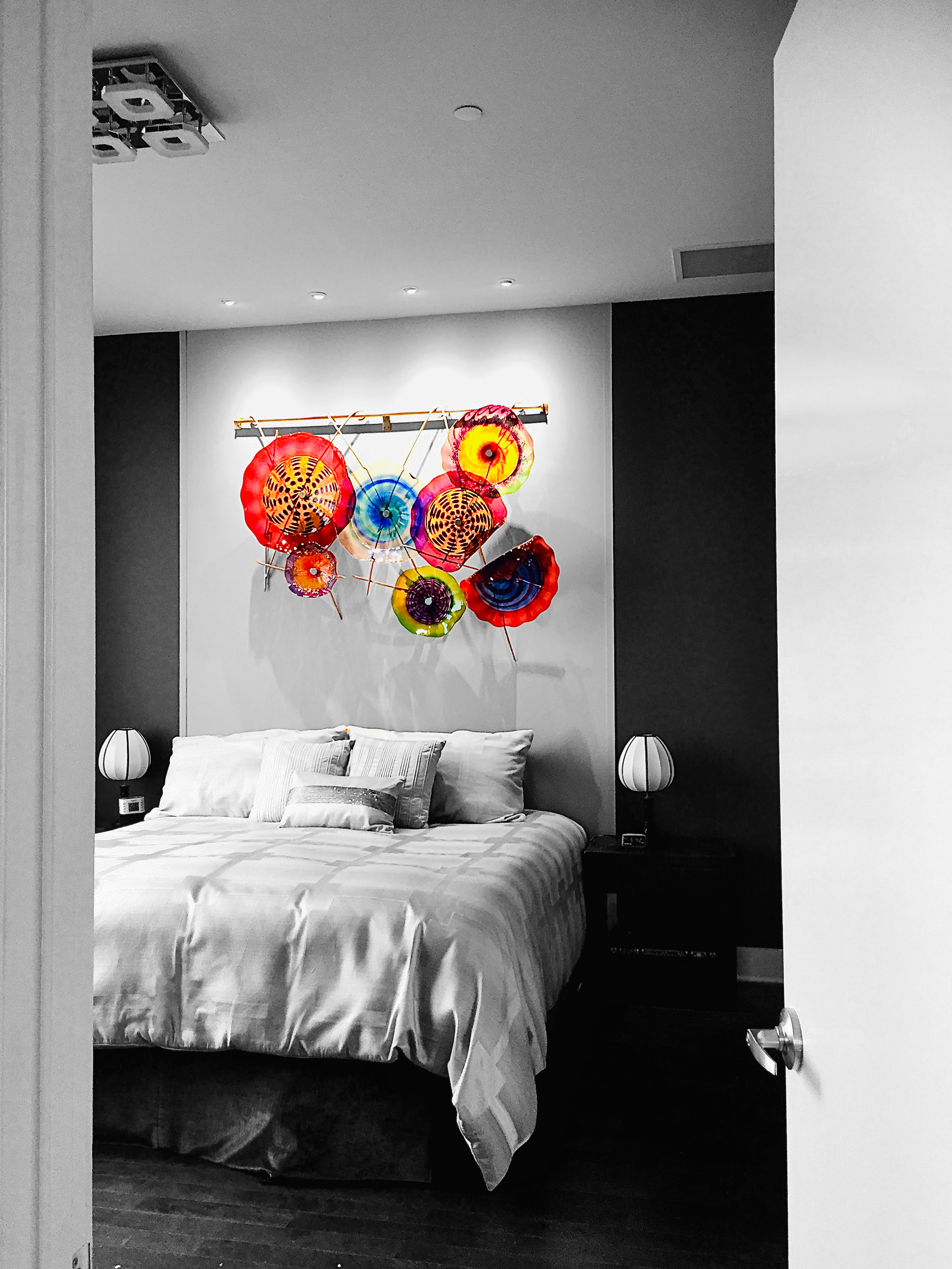 A modern bedroom with a black and white color scheme, featuring a bed with white bedding, two side tables with white lamps, and colorful umbrella-inspired art on the wall.