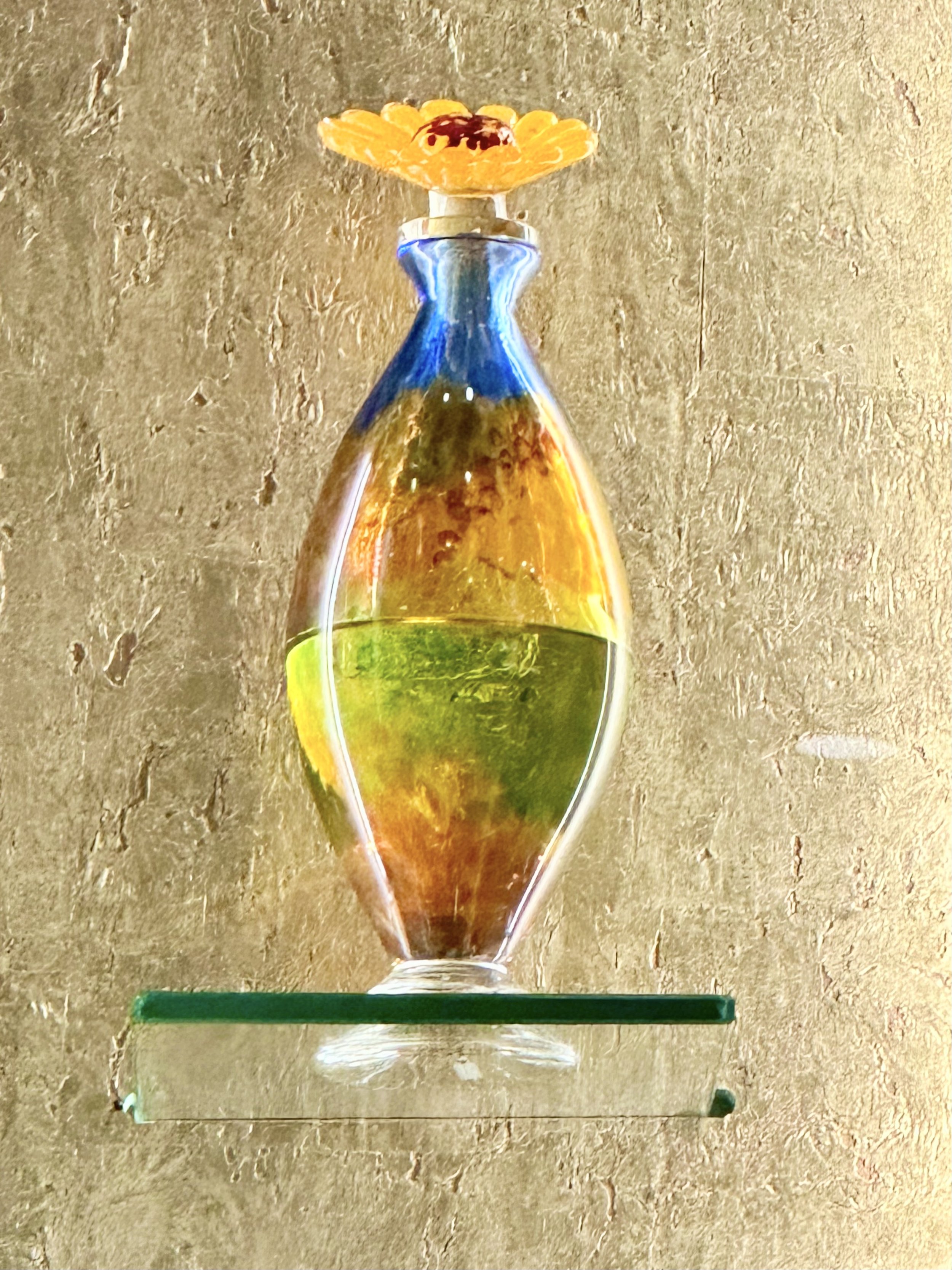 Colorful glass sculpture of a perfume bottle with a flower on top, displayed on a glass shelf against a textured background.
