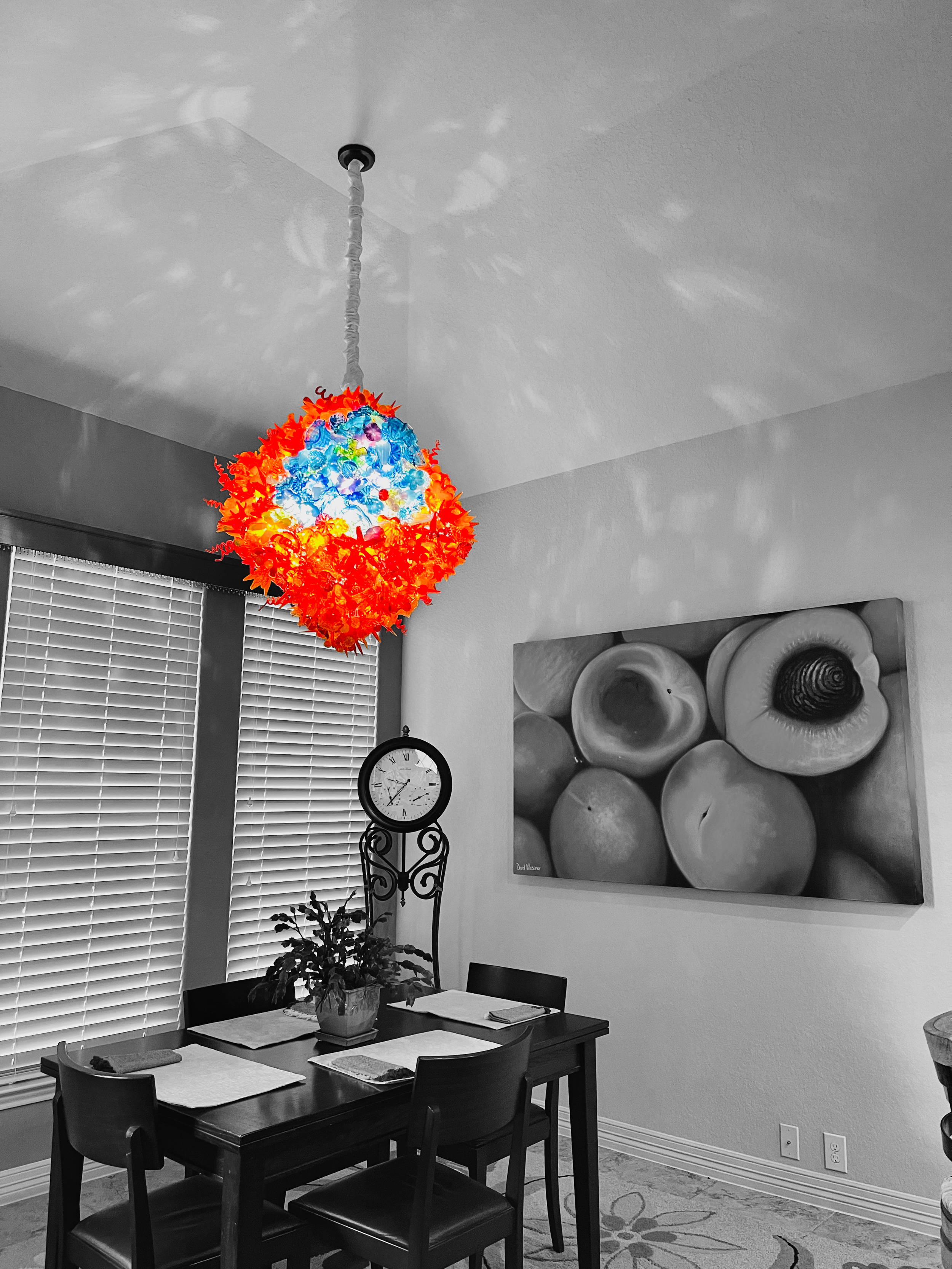 Colorful paper lantern hanging from ceiling in a mostly black and white dining room with a table, four chairs, potted plant, wall clock, window with blinds, and a fruit-themed painting on the wall.