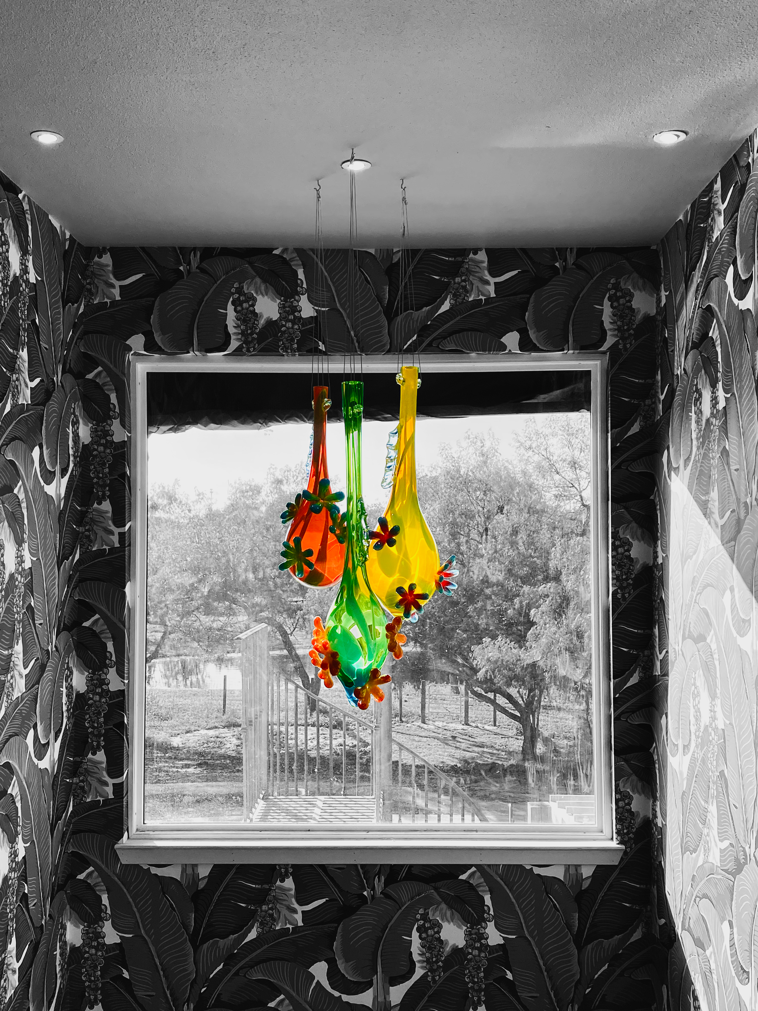 Colorful glass pendant light hanging in front of a window with black and white landscape outside, surrounded by dark leafy patterned wallpaper.