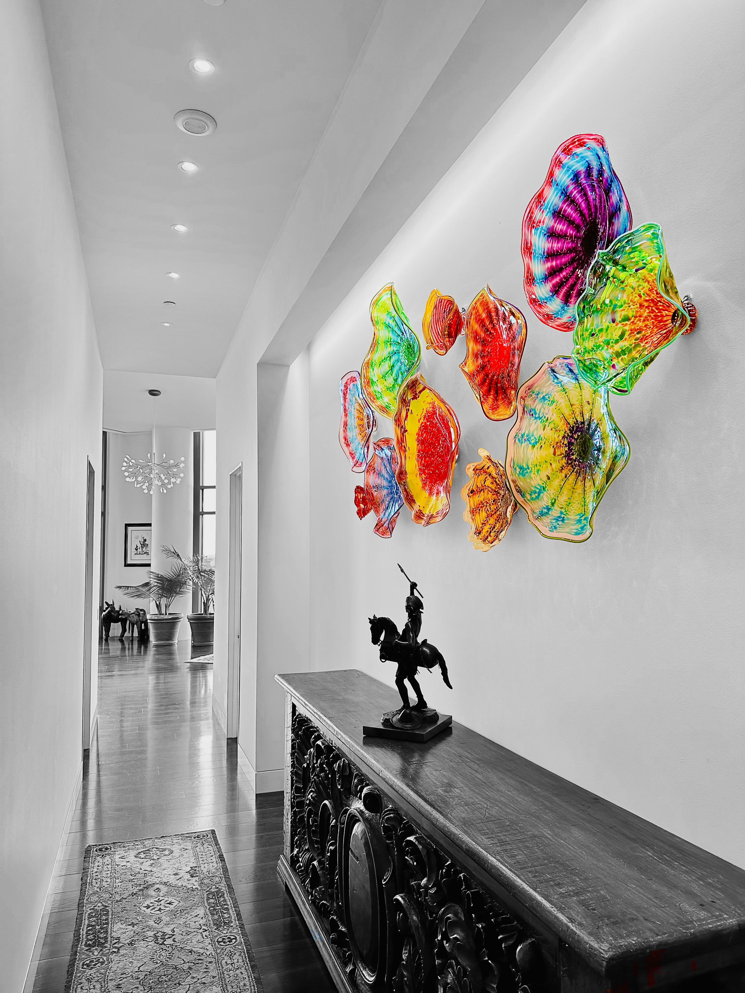Colorful glass art installation on a white wall in a hallway with a wooden console table and a black sculpture of a mounted knight. The hallway has a patterned rug and leads to a room with potted plants and a chandelier.
