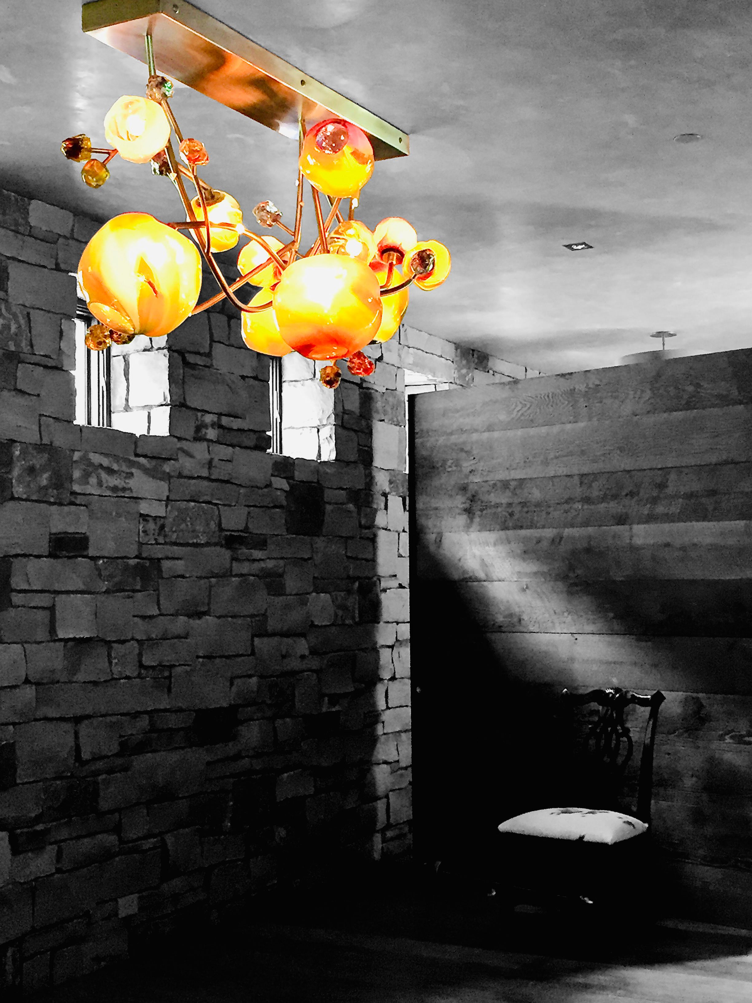 A bright orange chandelier hanging from a ceiling in a dimly lit room with stone brick walls and a wooden wall, with a chair with a white cushion visible in the background.