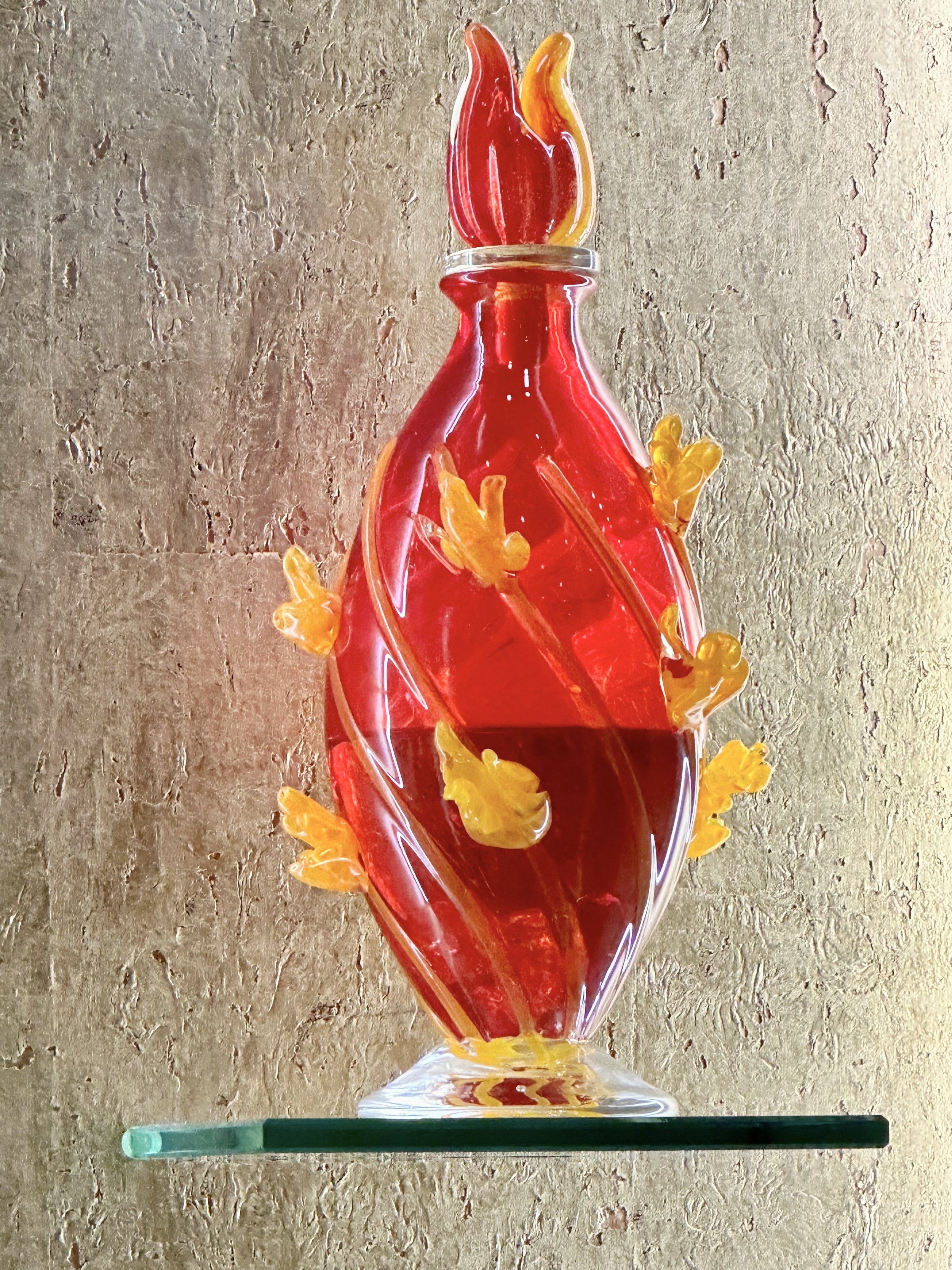 A decorative glass sculpture resembling a flaming torch with red, yellow, and orange glass flames and a red and yellow body, mounted on a clear glass base.