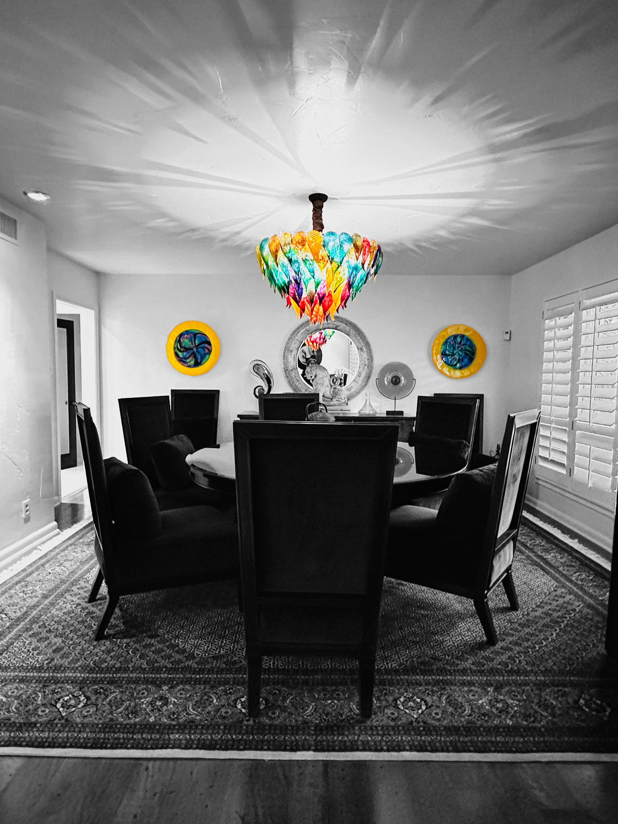 A dining room with a colorful, rainbow glass chandelier hanging from the ceiling, and a black and white color scheme for the walls and dining furniture. The room features a round dining table with six black chairs and decorative wall art, including two circular stained glass pieces and a large mirror.