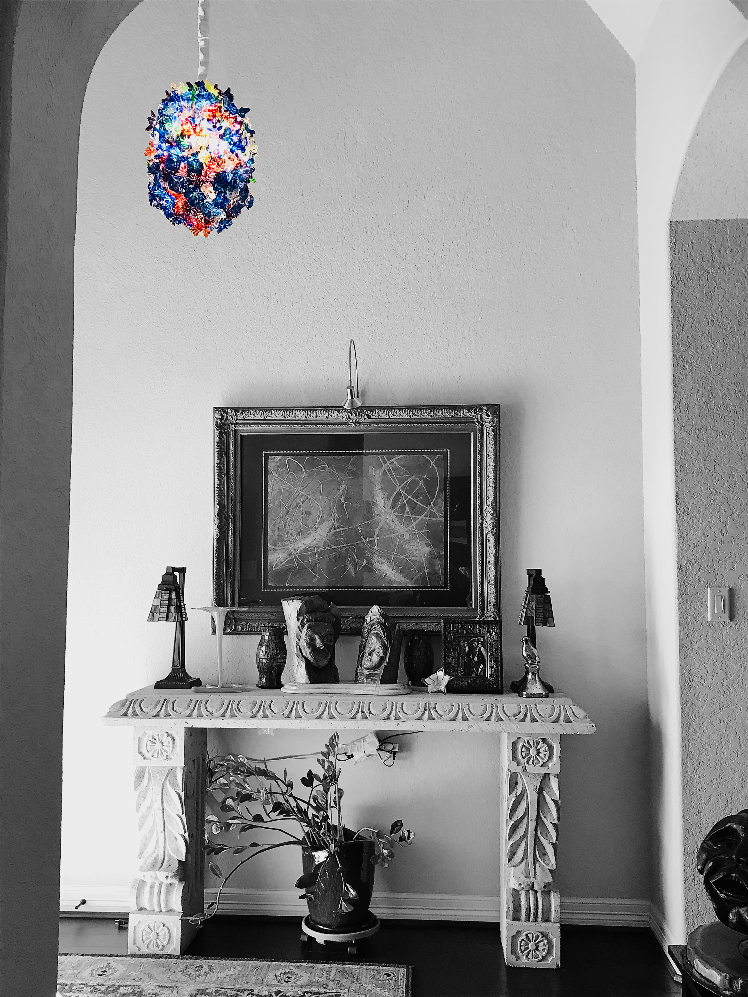 Black and white photo of a decorated fireplace mantle with sculptures, a framed mirror or artwork, and two small table lamps. There is a potted plant on the floor in front of the mantle. A colorful hanging sculpture is visible in the upper left corner, contrasting with the black-and-white scene.