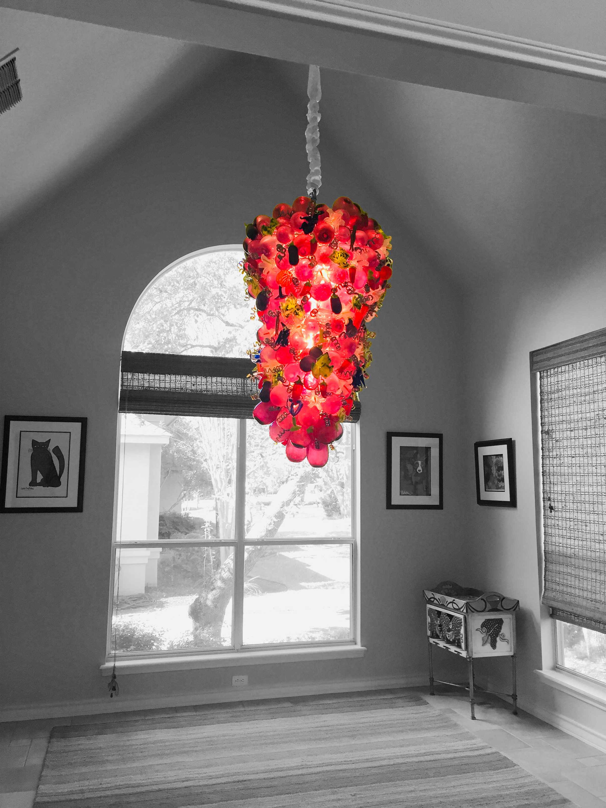 Colorful, artistic chandelier hanging from a ceiling in a room with black and white decor, featuring framed pictures on the wall and a window showing an outdoor scene.