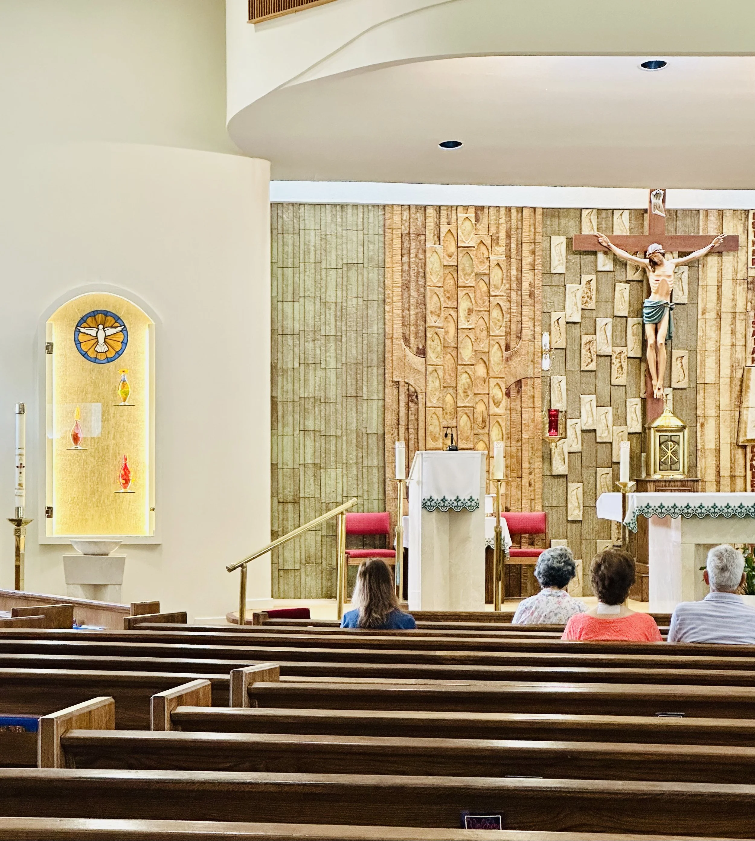 Inside a church with a crucifix on a decorated wooden wall, three people sitting in pews, and an altar with candles and a cloth.
