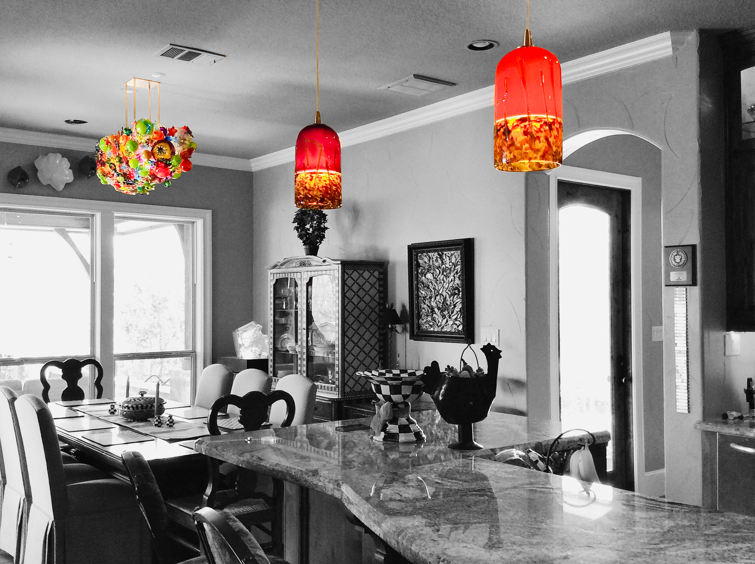 Colorful pendant lights hanging over a kitchen island in a dining area, with some furniture and decorations in the background, in an otherwise black and white photo.