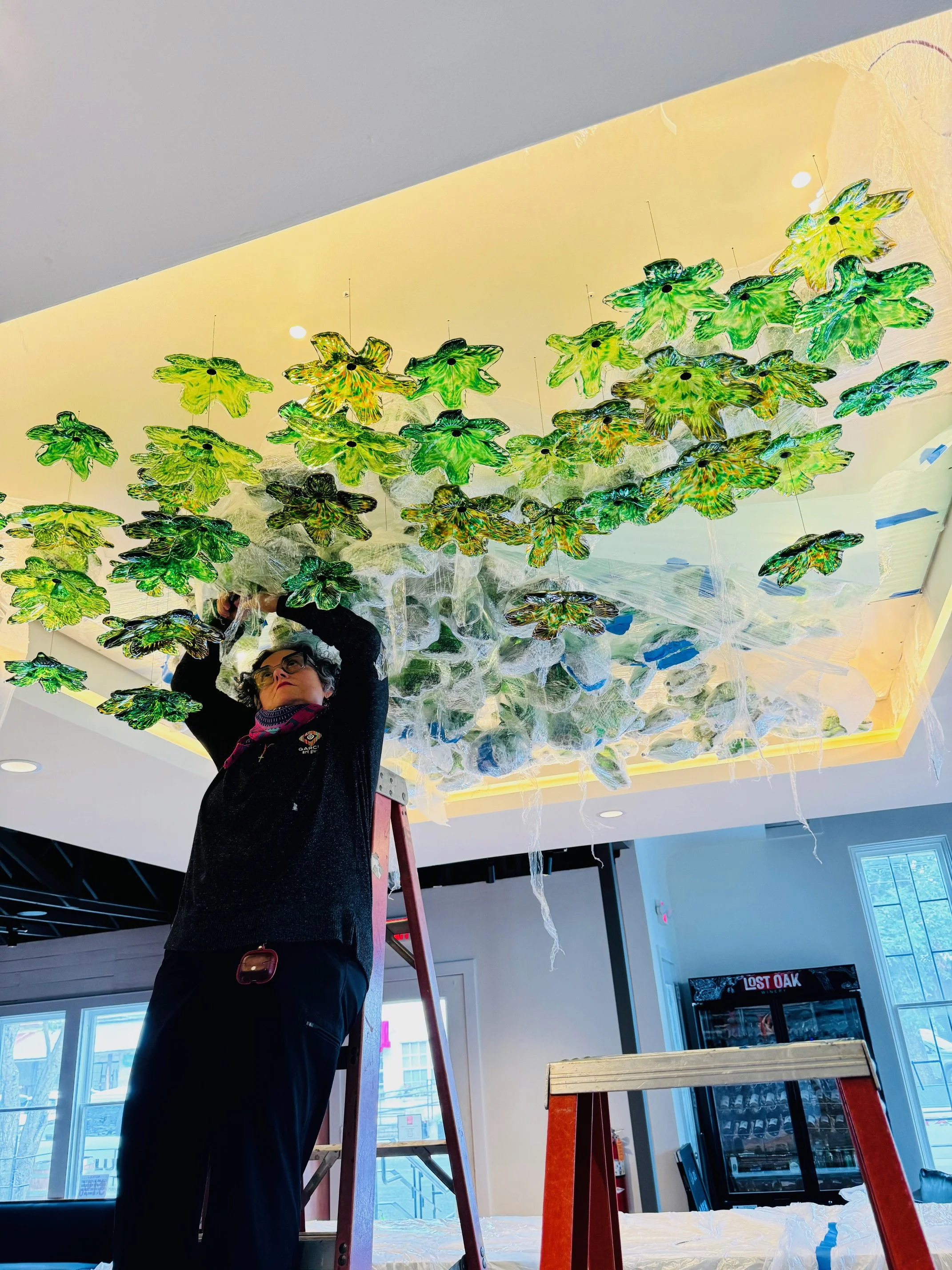 Person standing on a ladder installing a ceiling light fixture with decorative green and black leaf-shaped objects in a commercial space.