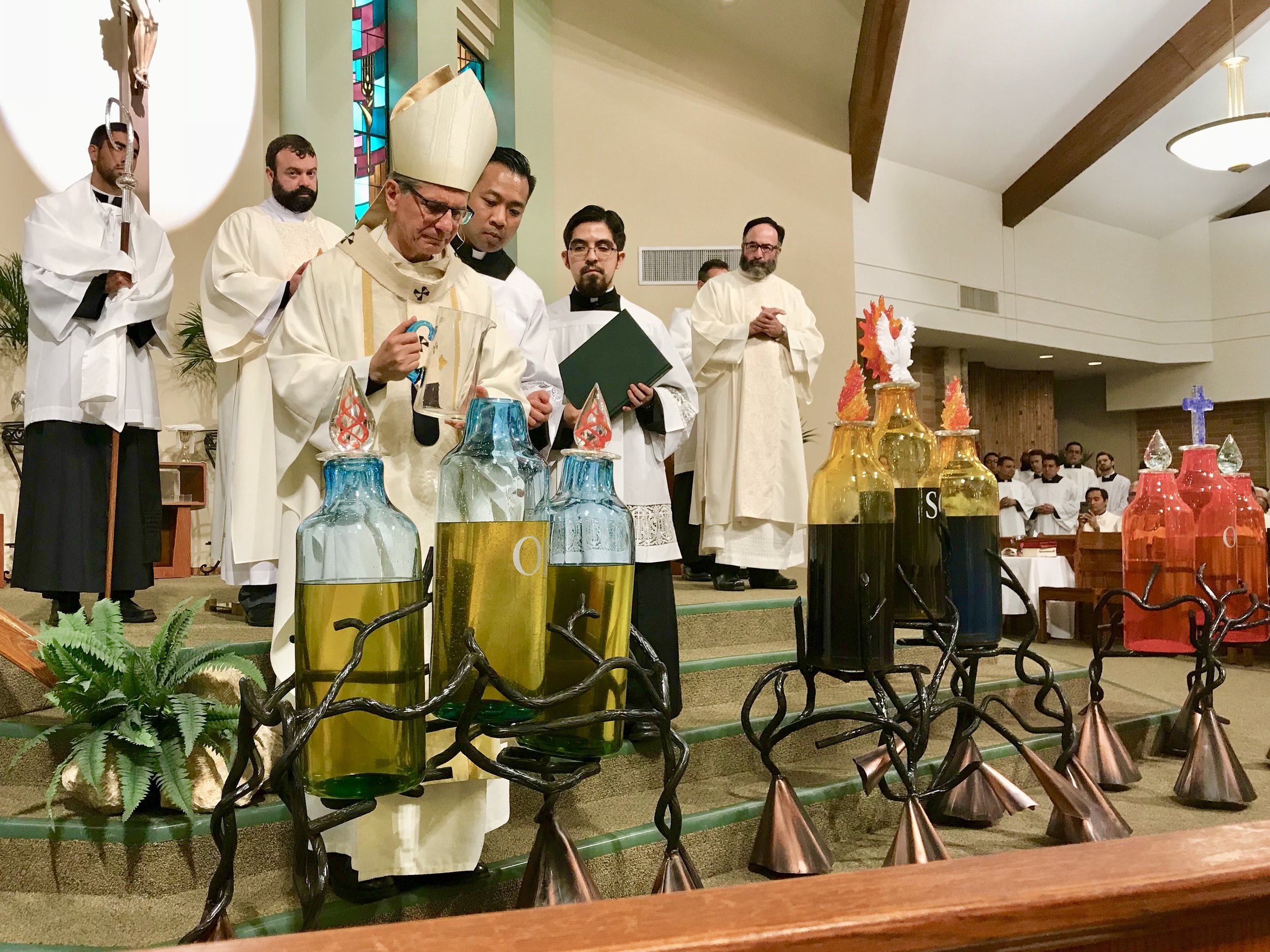 Catholic priests and altar servers standing on the altar during a religious ceremony, with large glass bottles containing colored liquids and decorative flames in front of them.