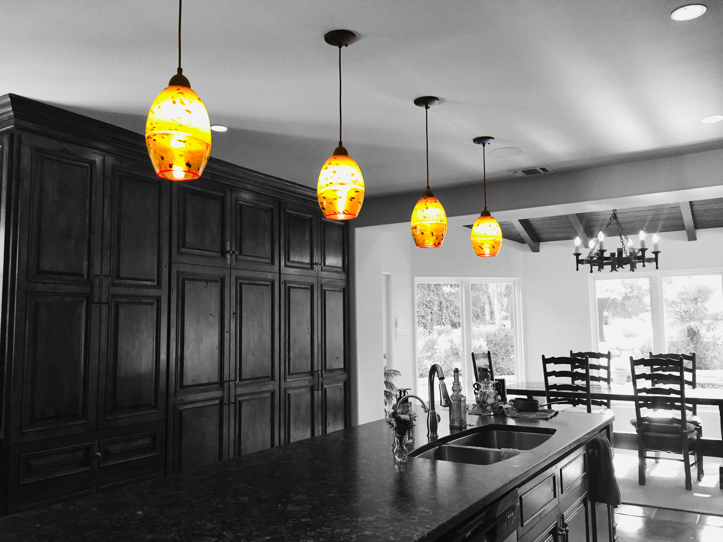 Colorful pendant lights hanging above a kitchen counter with black cabinets and a dining area with a chandelier and large windows.