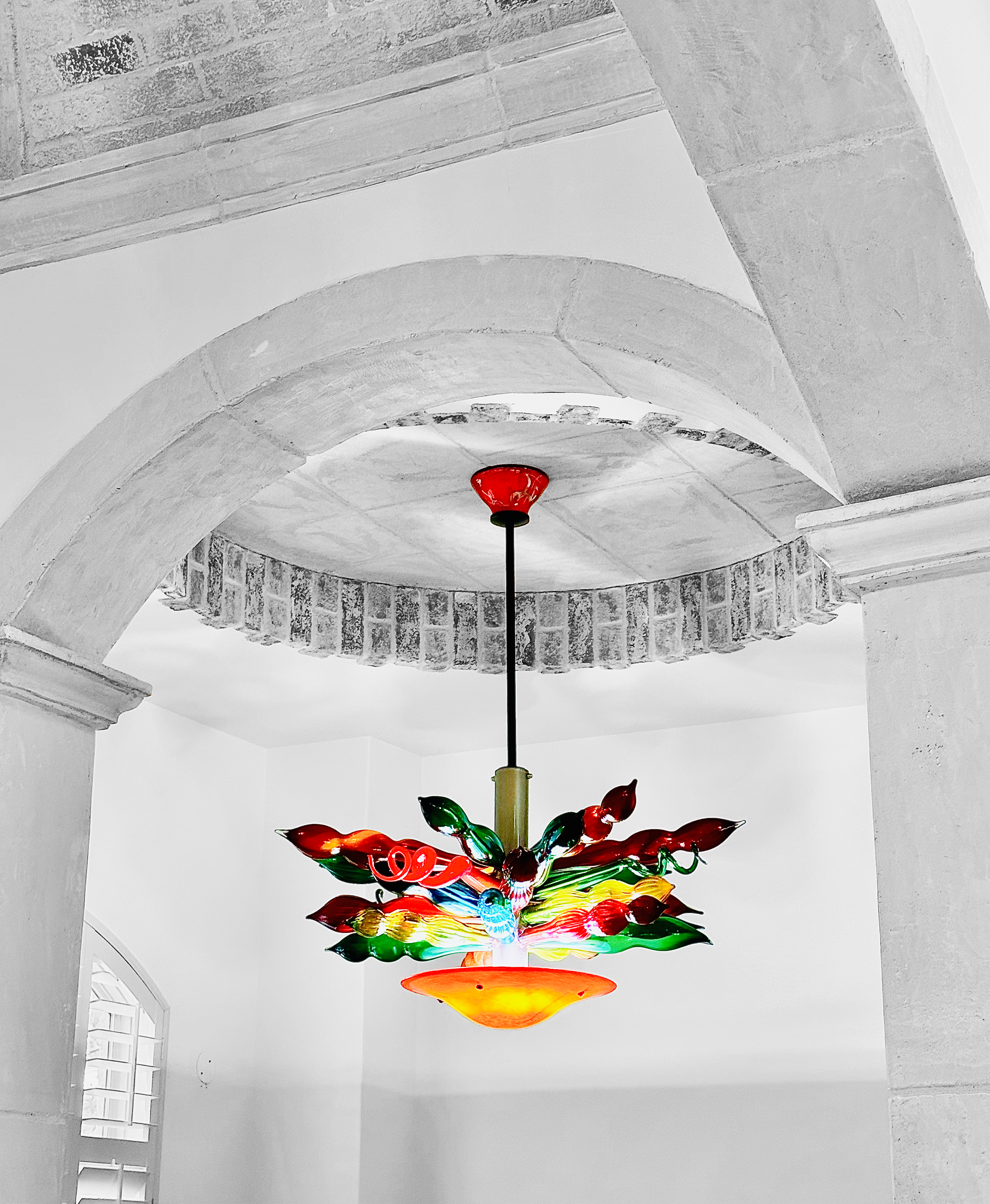 Colorful glass chandelier with red, green, yellow, and blue glass elements hanging from a black cord in a white room with a curved brick ceiling and white walls.