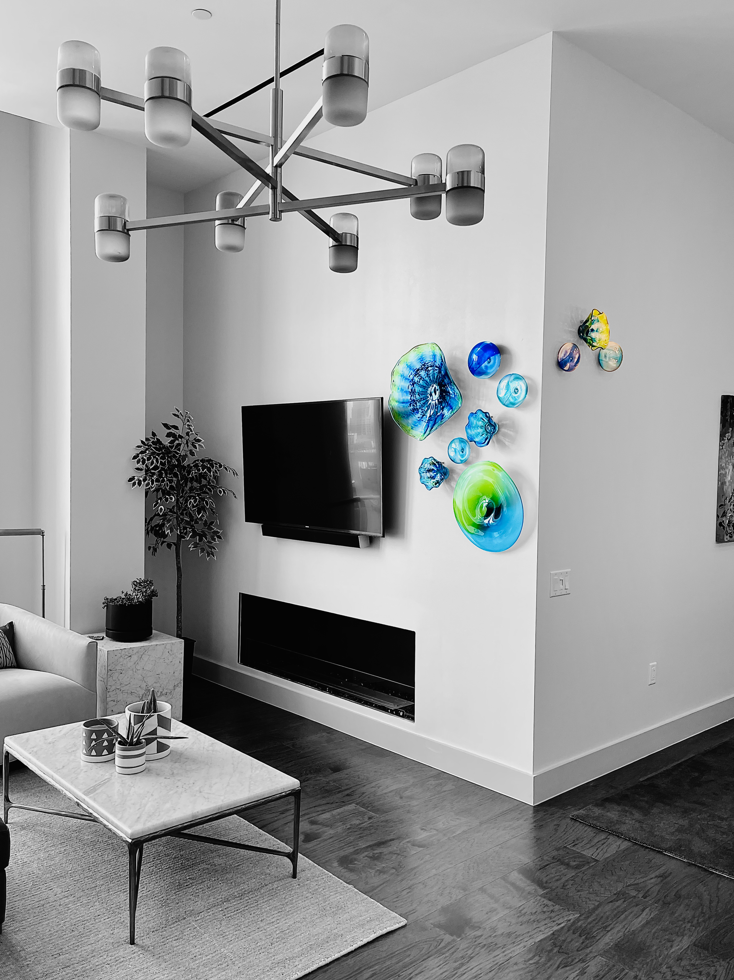 Modern living room with a white wall decorated with colorful glass art, a flat-screen TV, a fireplace, a marble coffee table, a beige sofa, a houseplant, and a contemporary chandelier.