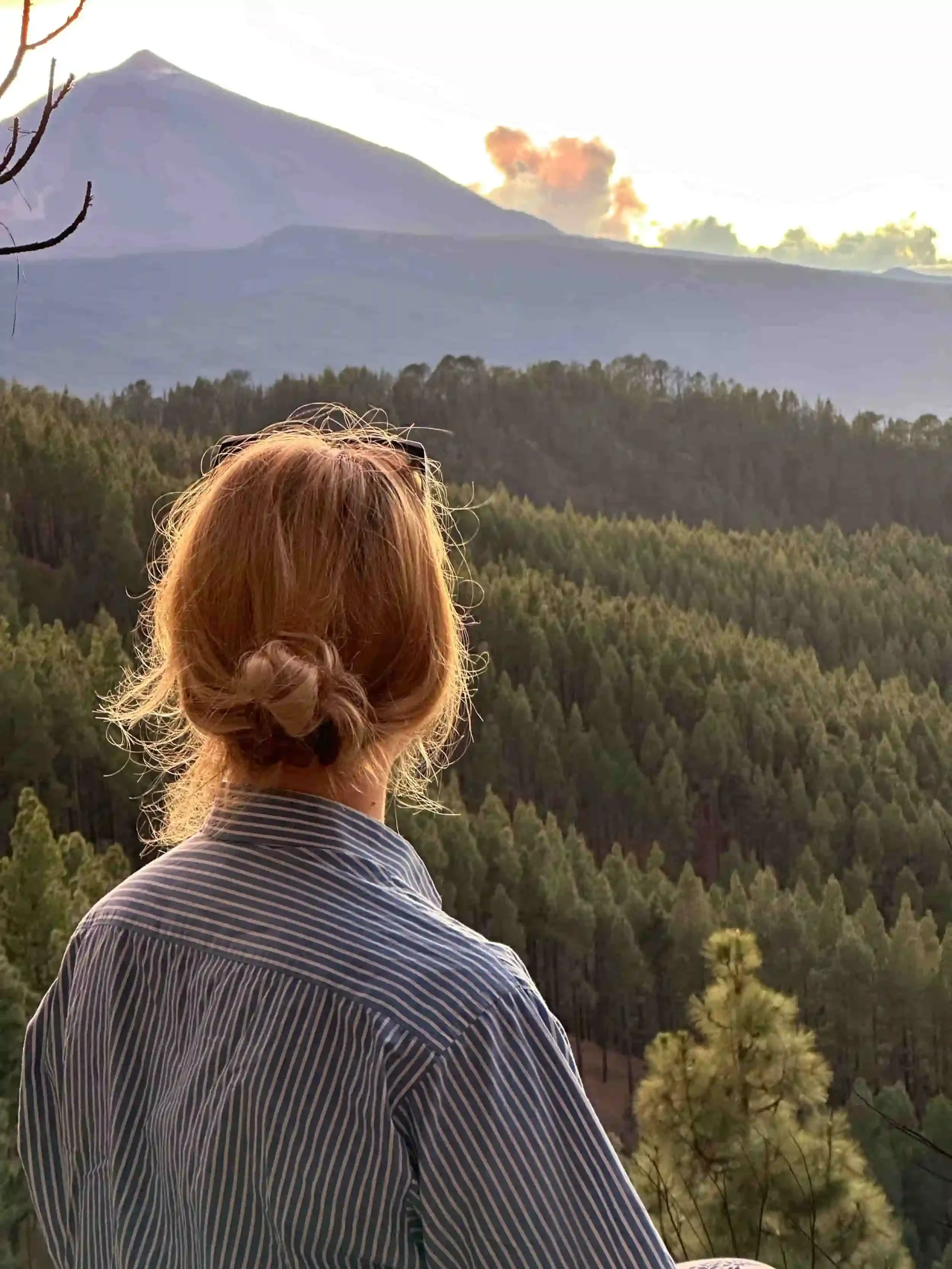 Woman with blonde hair in a bun, wearing a striped shirt, looking at a forested mountain landscape with a volcanic mountain in the background during sunset.