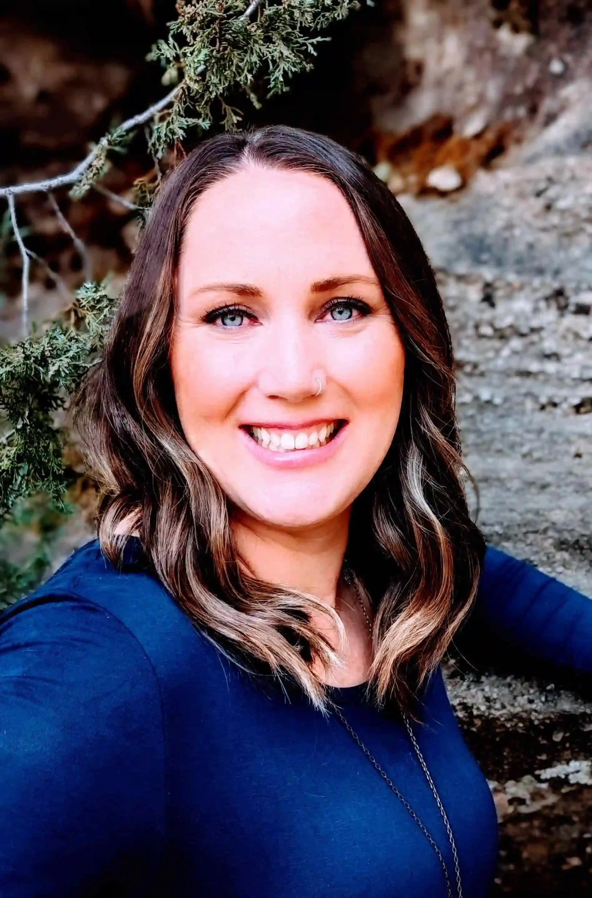 A smiling woman with wavy brown hair and blue eyes, wearing a navy blue top, outdoors near a rocky area and green foliage.