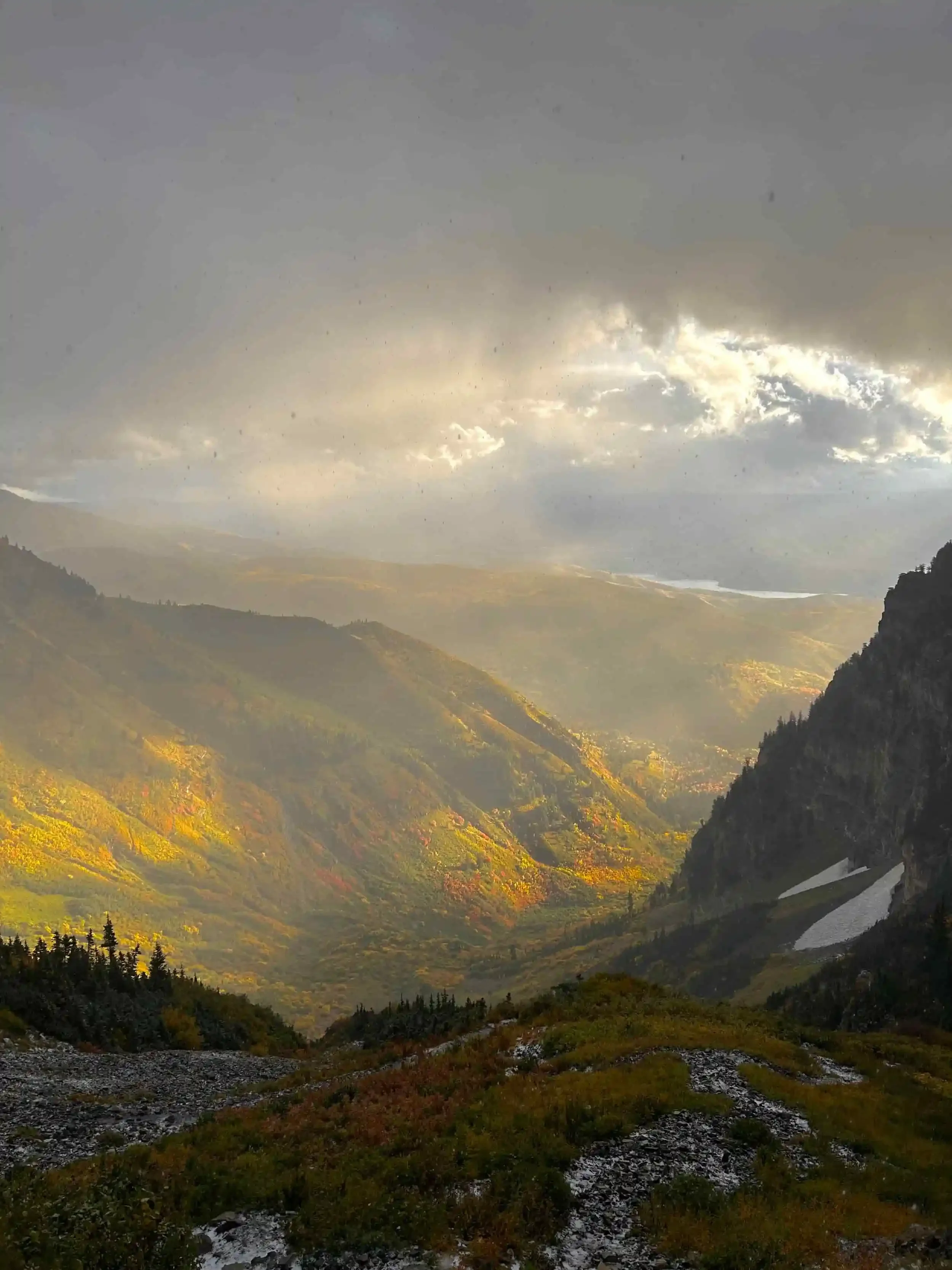 A mountainous landscape with a cloudy sky, sunlight breaking through, featuring green and yellow foliage, rocky terrain, and patches of snow on the mountains.