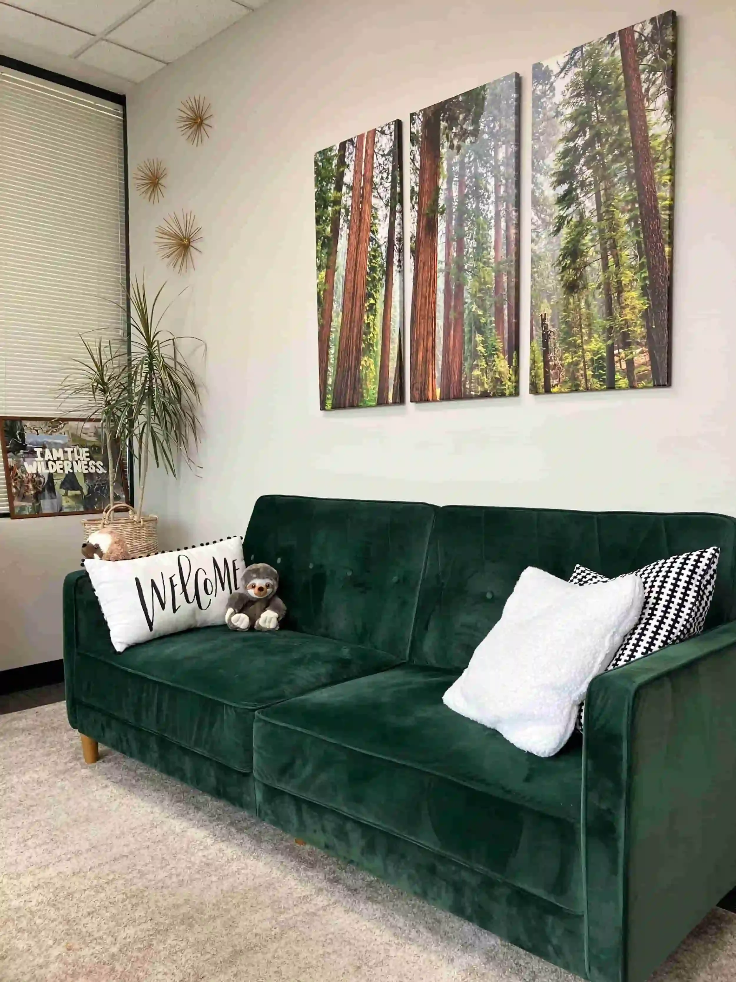 A cozy green velvet sofa with plush pillows and a stuffed animal in a therapy office with framed forest landscape art and decorative wall accents.