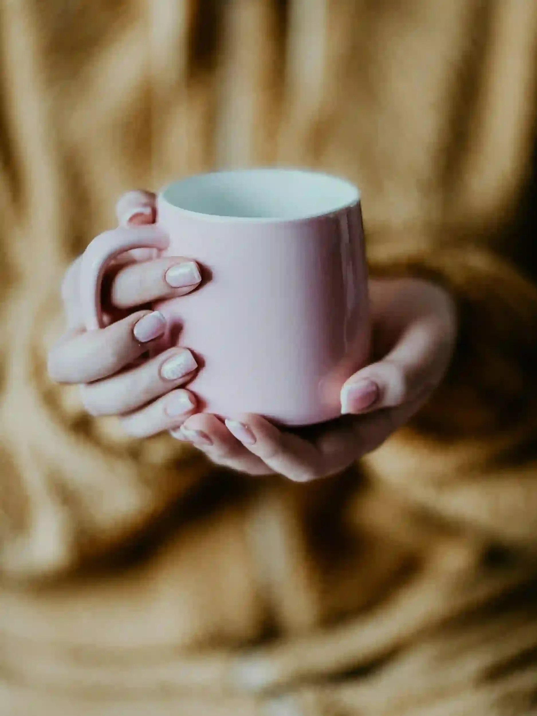 A person with manicured nails holds a pink mug in both hands, wearing a brown, fuzzy sweater.