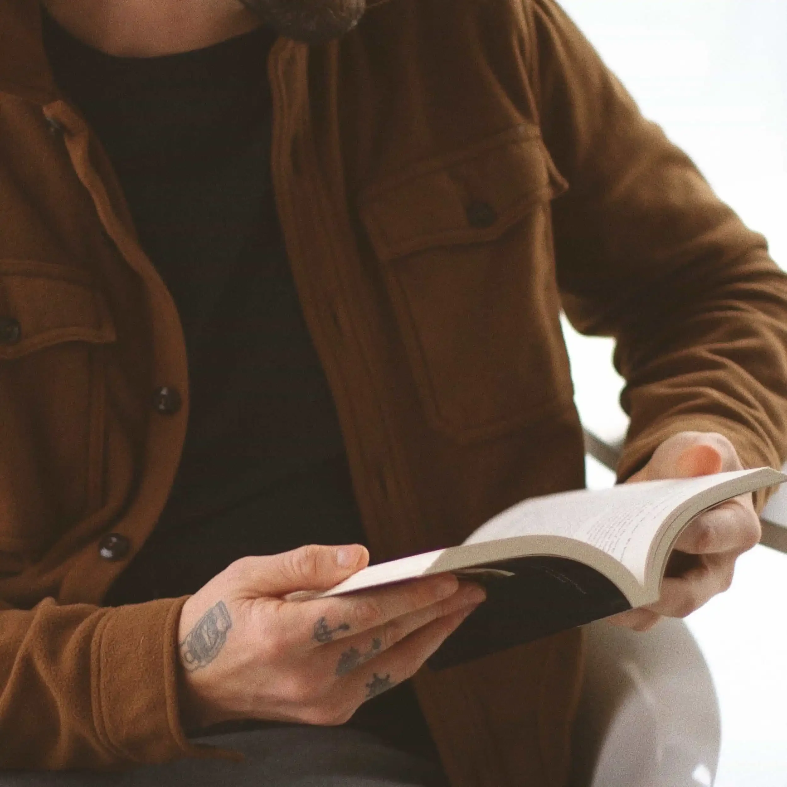 A person wearing a brown jacket and black shirt is sitting and reading a book. Their hands, which have small tattoos, are holding the open book. The background is softly lit and out of focus.