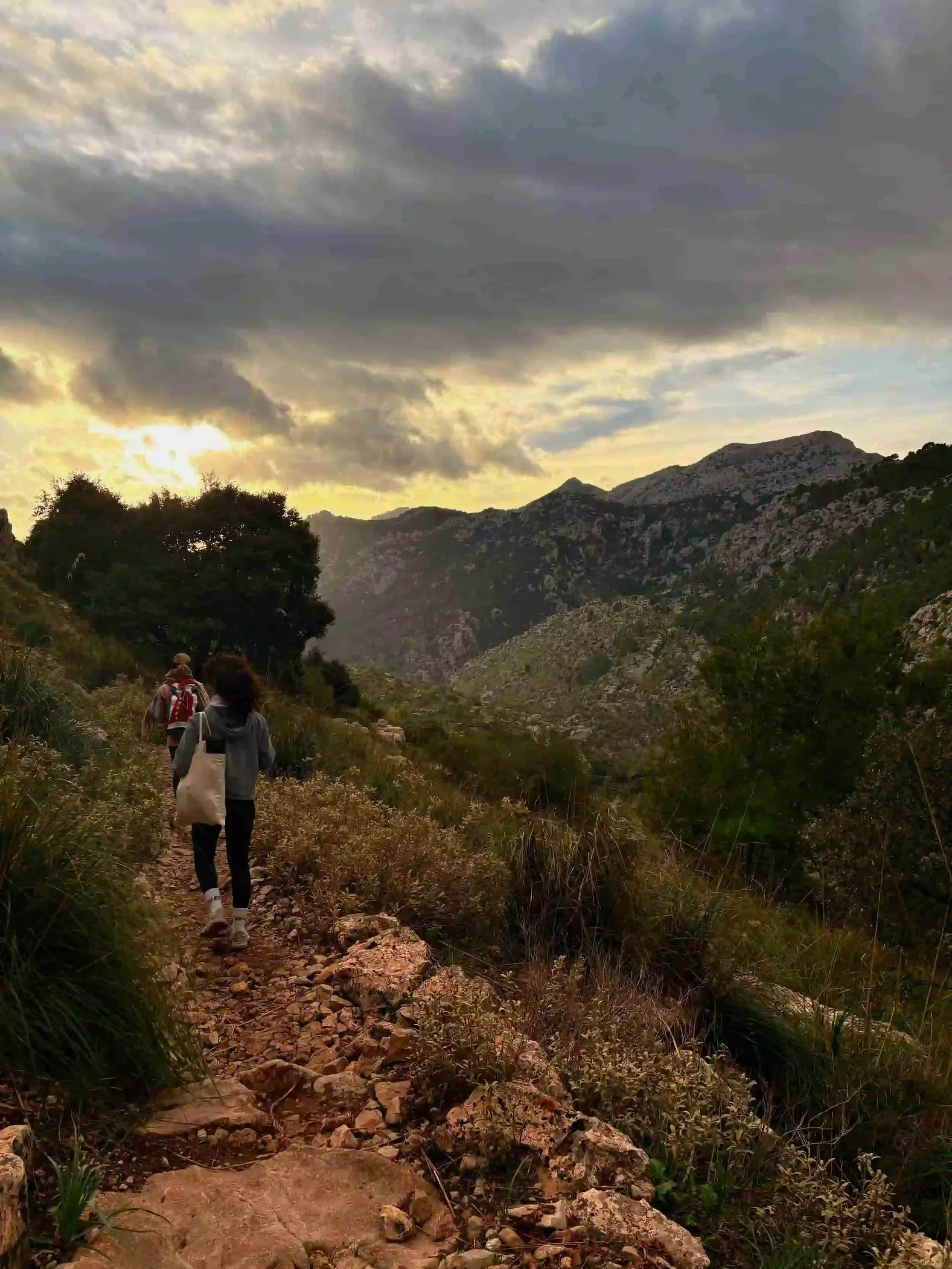 People hike on a rocky trail through a mountainous landscape under a cloudy sky during sunset.