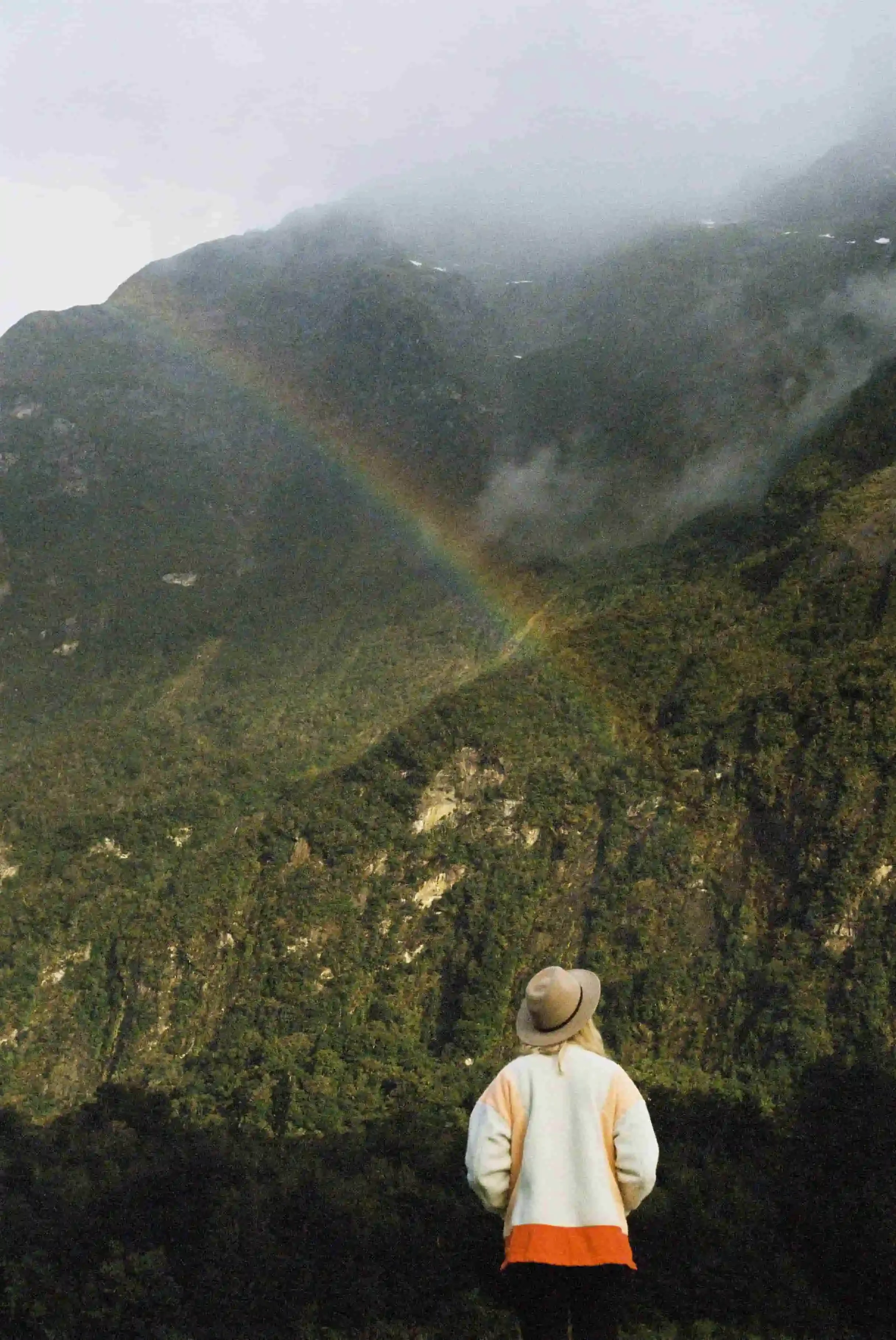 A person wearing a beige hat and colorful jacket looking at a rainbow over a lush green mountain with clouds and mist.