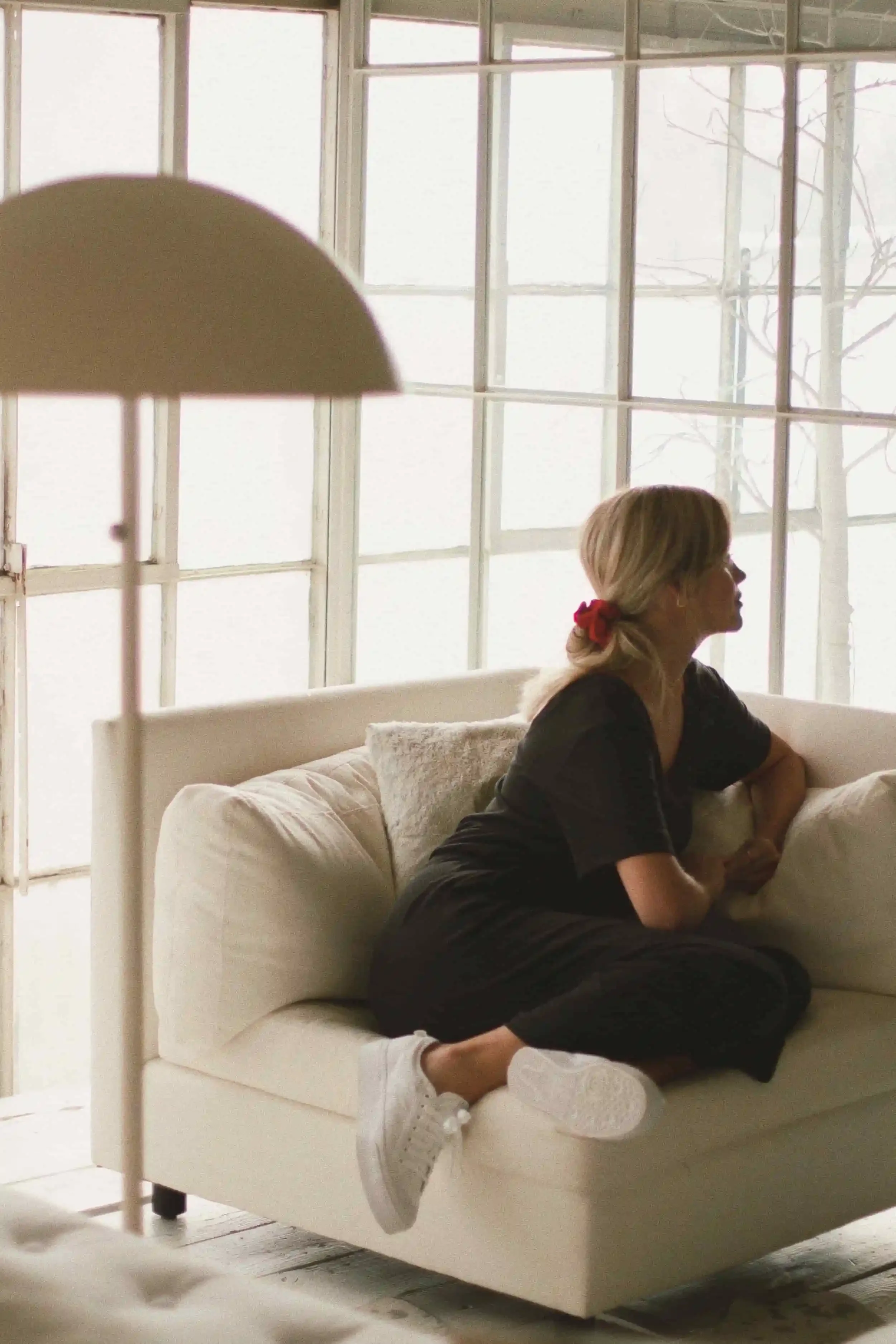 A woman with blond hair and a red hair tie sits sideways on a white sofa, looking out large, bright windows. She wears a black outfit and white sneakers. A modern white floor lamp stands in the foreground.