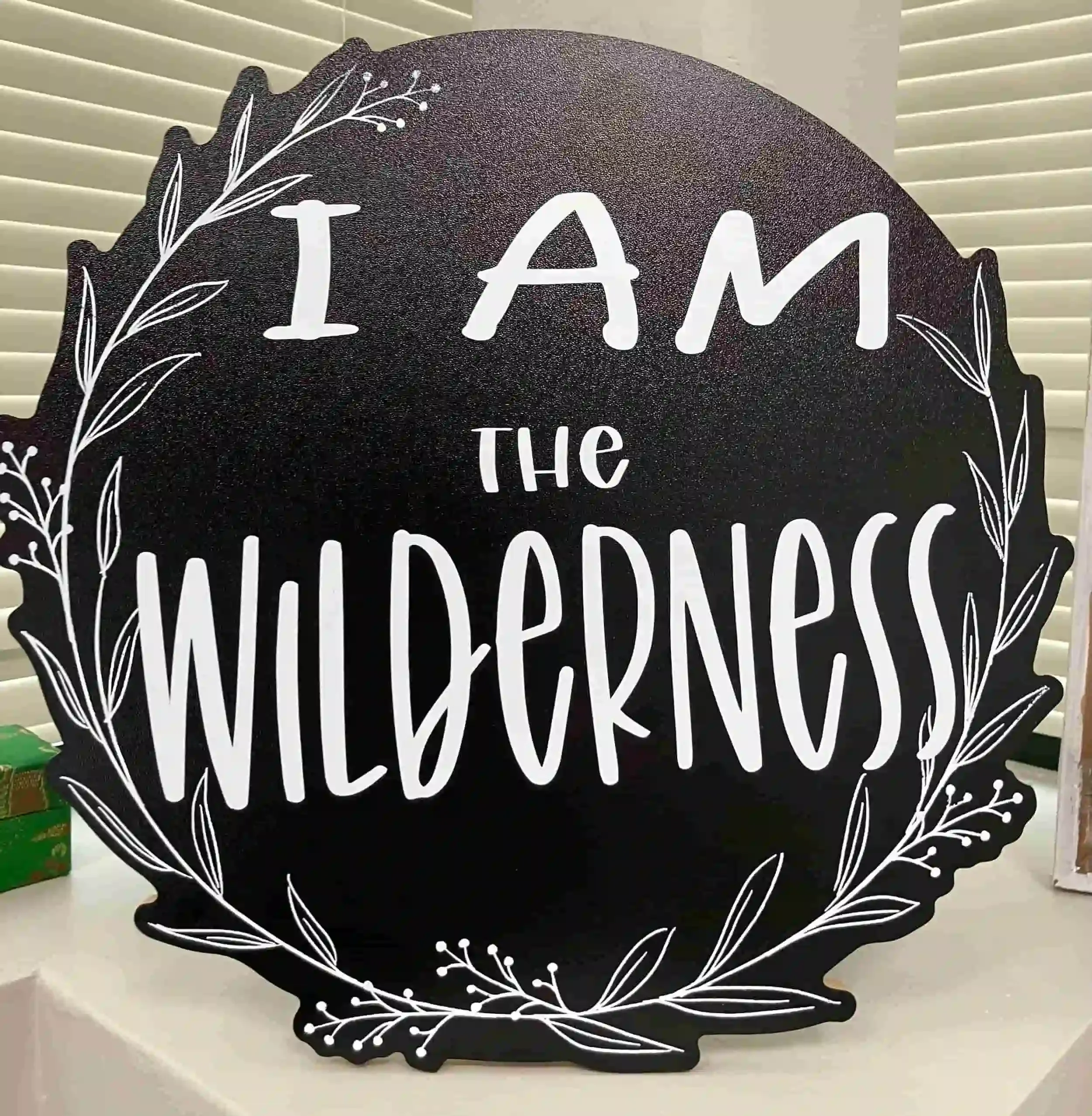 Decorative black circular sign that says "I am the wildness" with white lettering and a leafy border design.