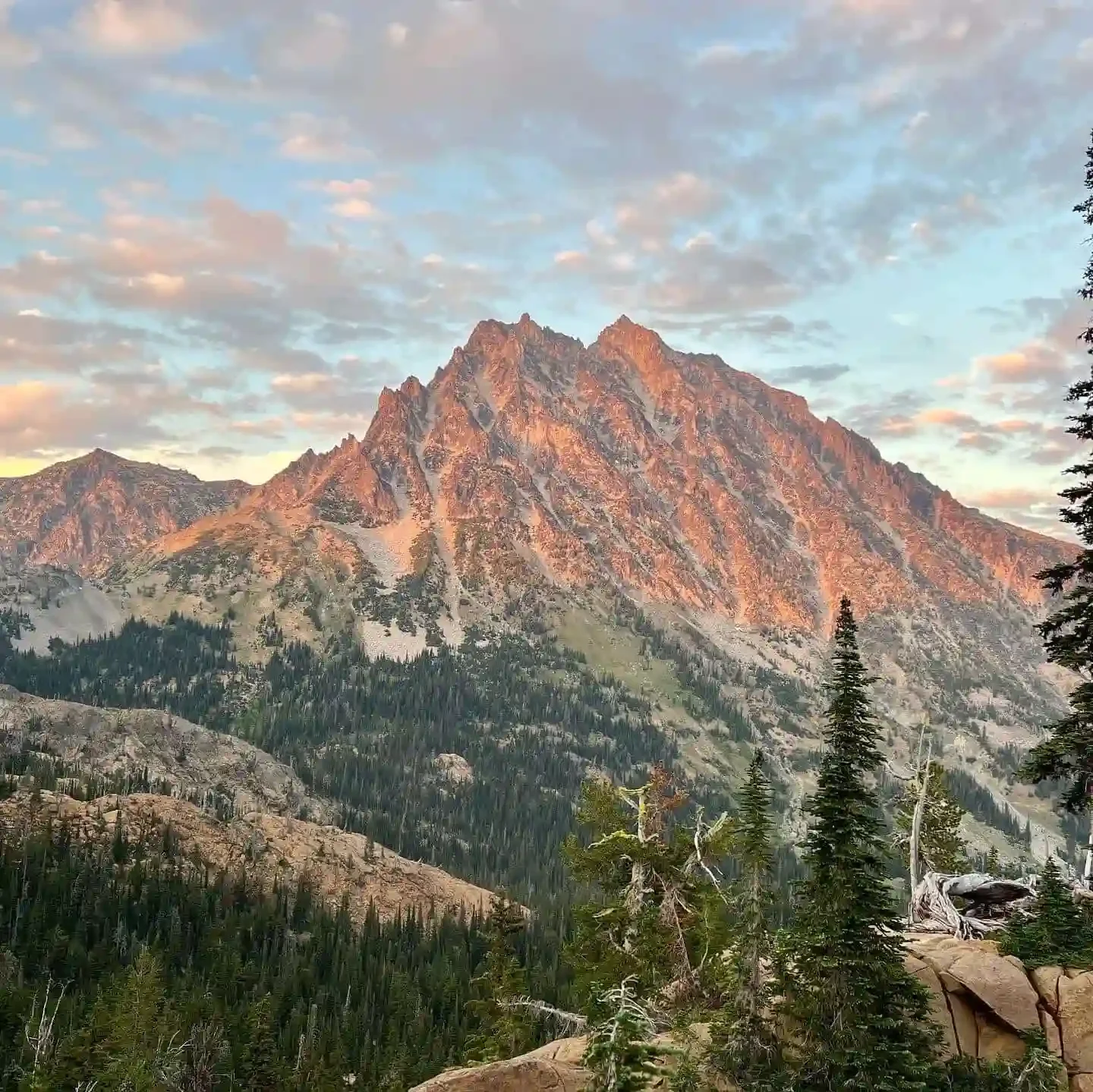 Sunset lighting on a rugged mountain peak with green forest at the base and a partly cloudy sky.