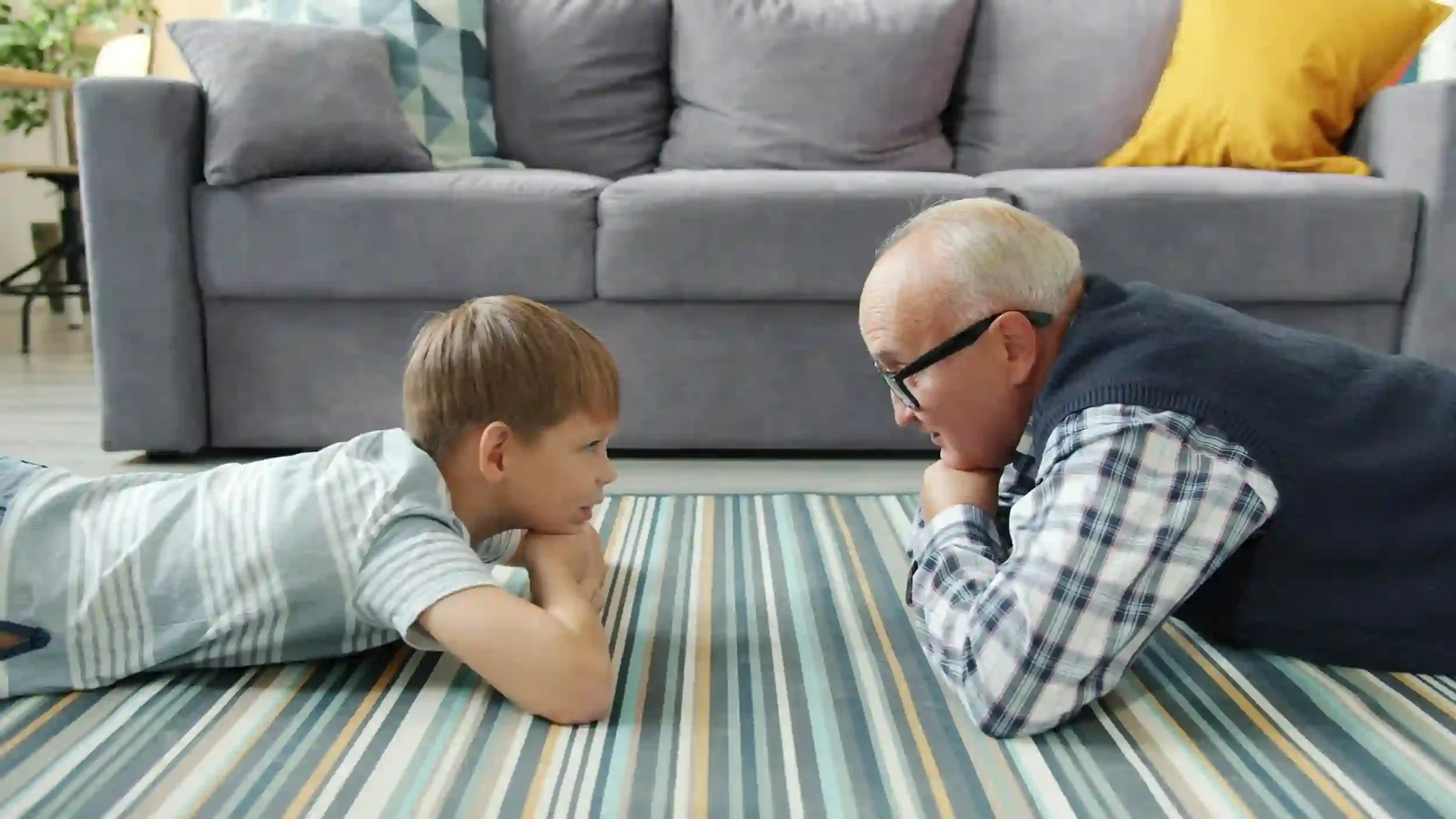 A boy and an older man lie on a striped rug facing each other, smiling, in a cozy living room with a gray couch behind them.