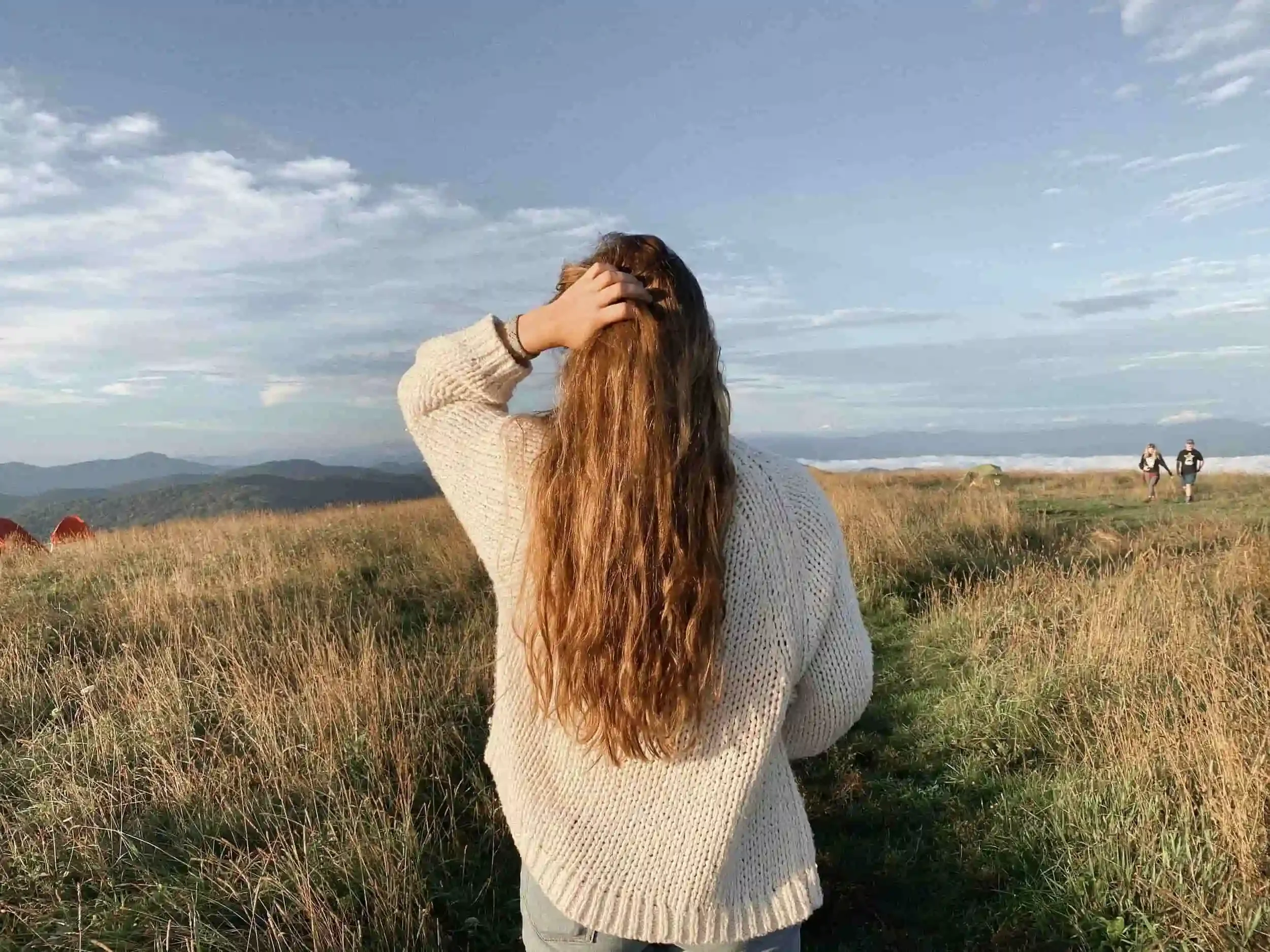 A woman with long, wavy hair wearing a cream-colored sweater standing on a grassy field, facing away, with her hand on her head. In the background, two people walk on a trail with mountains and a partly cloudy sky overhead.