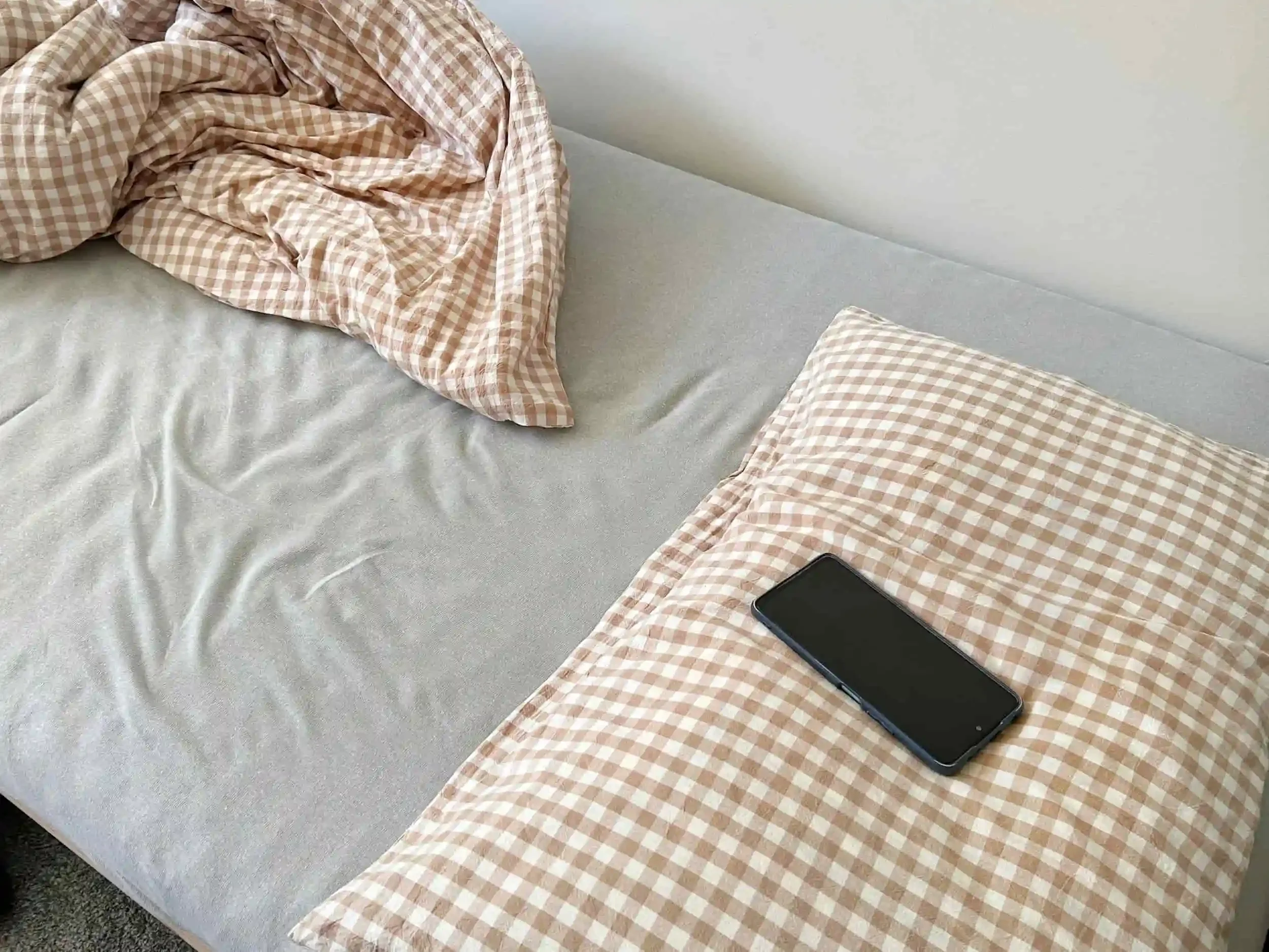 A smartphone rests on a checkered pillow atop an unmade bed with a matching blanket and gray fitted sheet.