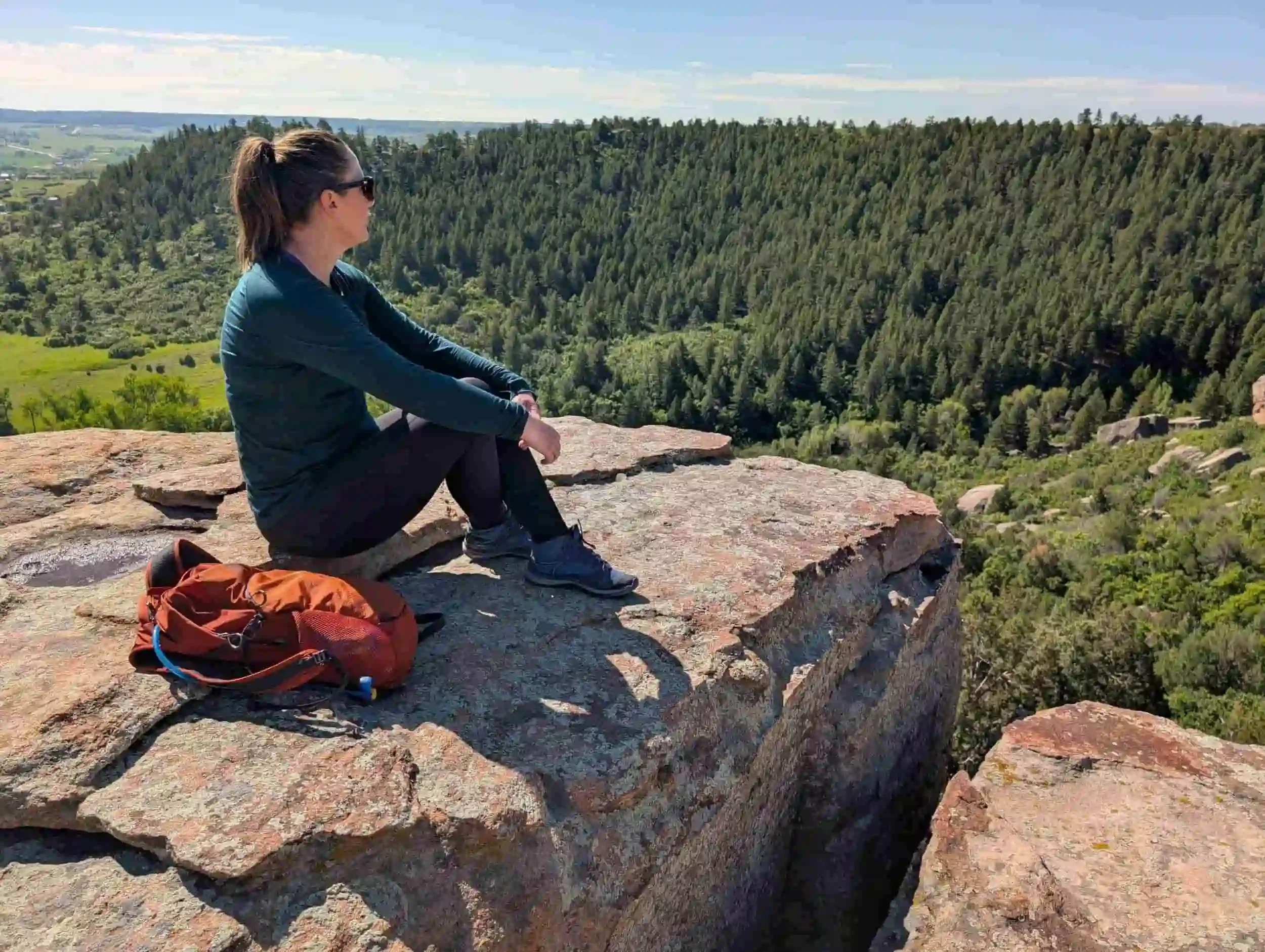 Alicia Rippy, EMDR psychotherapist, sits on a rocky cliff edge, looking out over a lush green forest and valley. She wears sunglasses, a dark jacket, leggings, and sneakers, with an orange backpack beside her. The sky is clear and sunny.