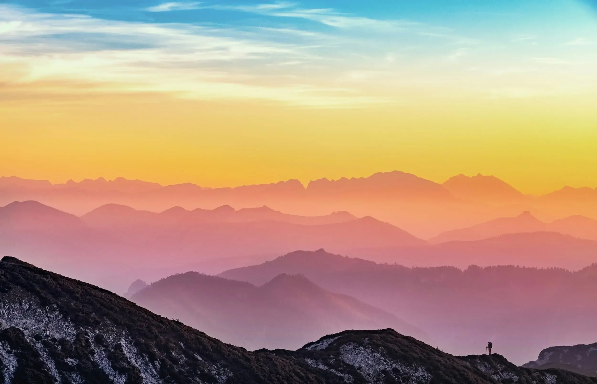 Colorful sunrise or sunset over layered mountain ranges with misty valleys, a clear sky transitioning from blue to orange and yellow, and a lone figure standing on a rocky foreground hill.