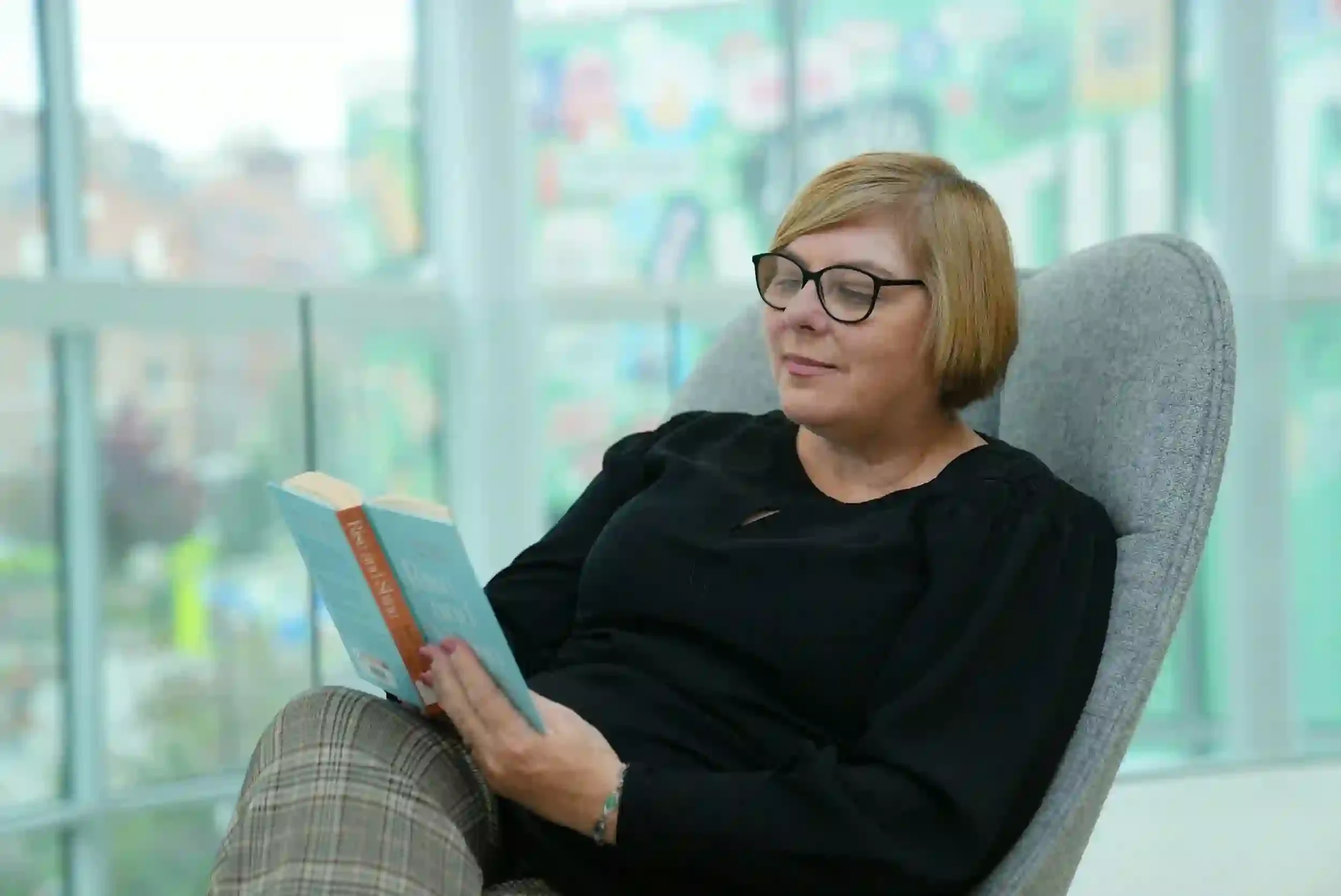 A woman with glasses sits in a gray chair, reading a book and smiling, with a bright, blurred background behind her.