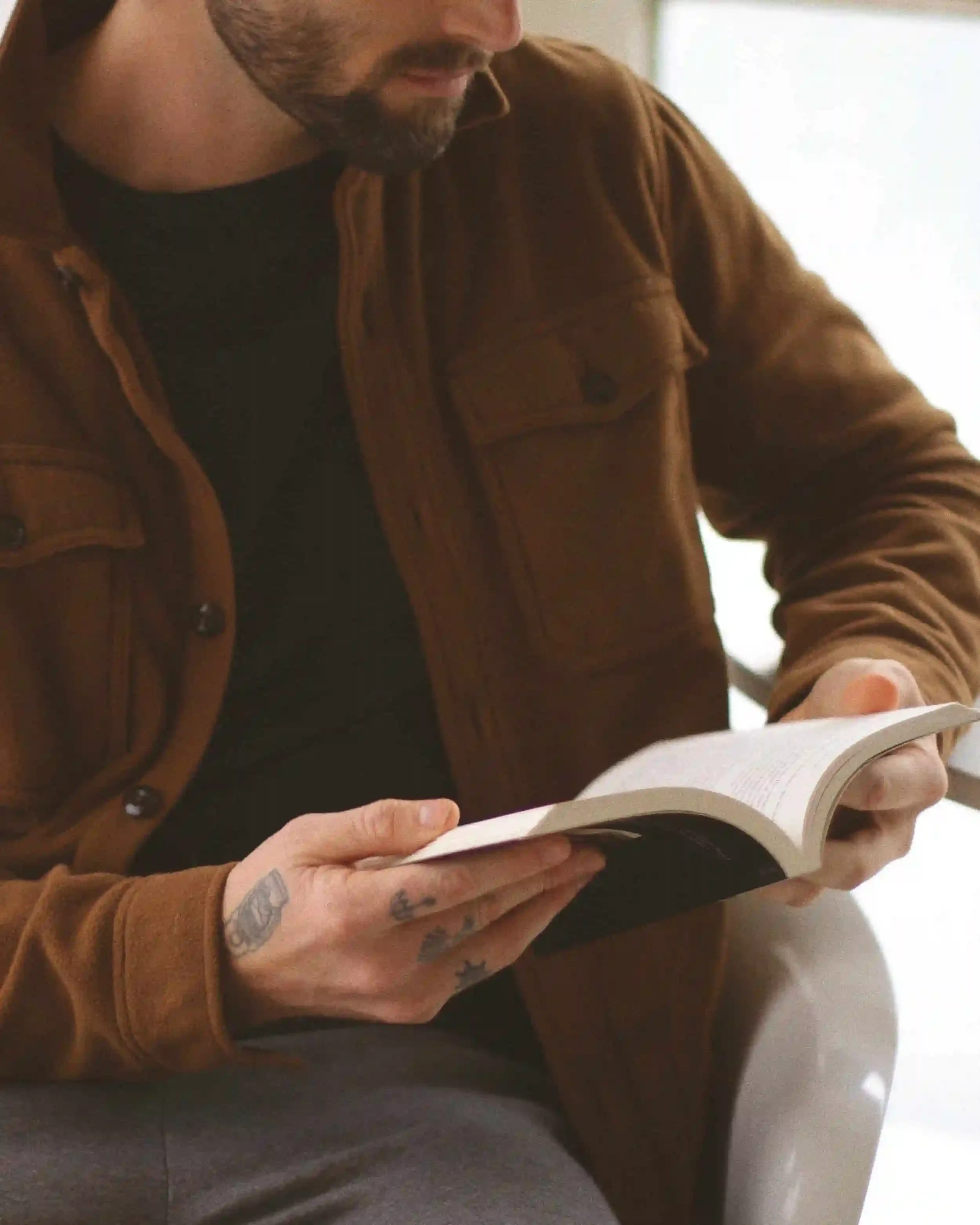 A man with tattoos on his fingers, wearing a brown jacket, is reading a book.