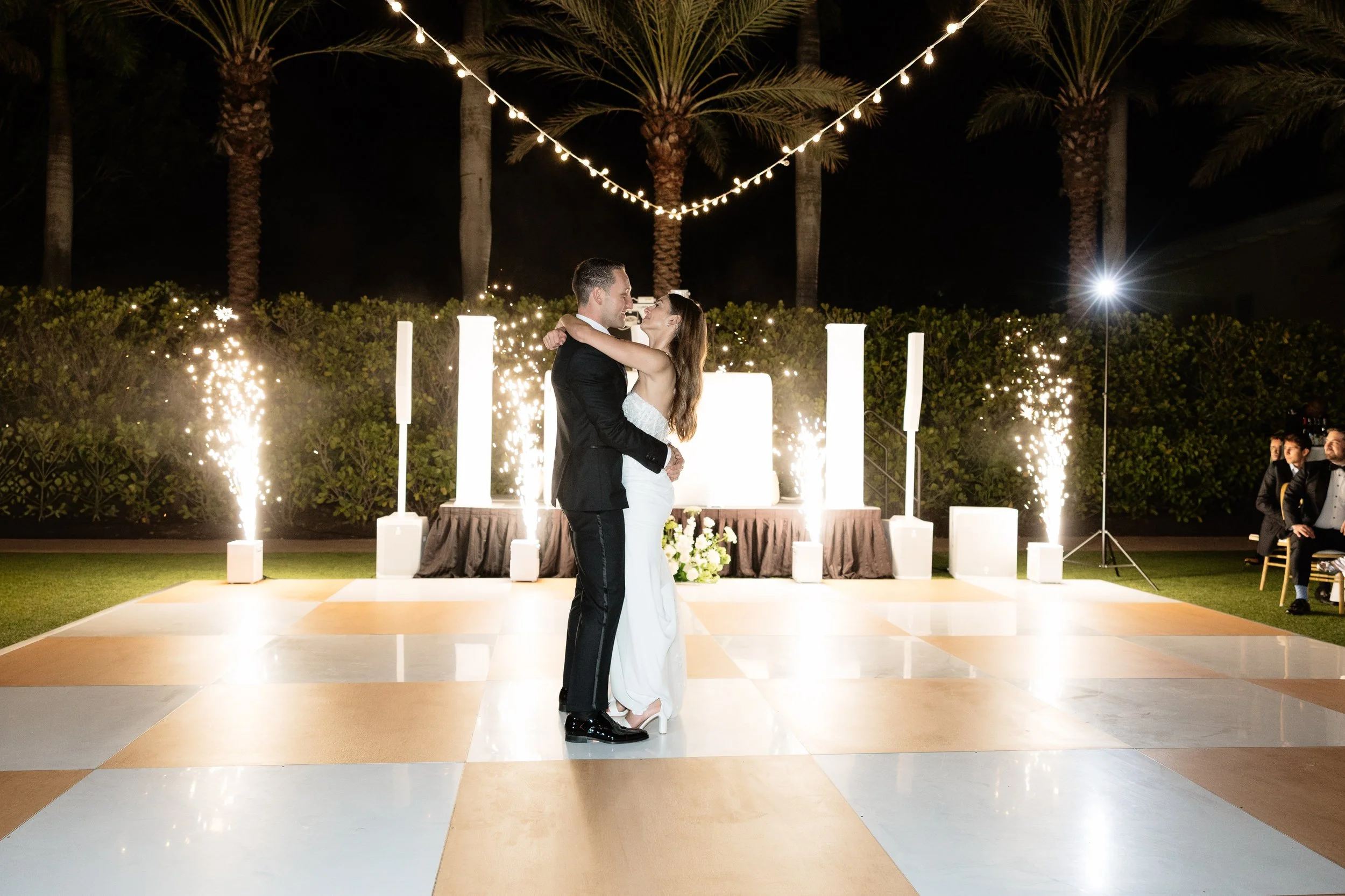 A bride and groom dance at their outdoor wedding reception at night, illuminated by string lights and sparklers, with palm trees in the background and guests seated on either side at the Hyatt Regency at Coconut Point in Bonita Springs, Florida 