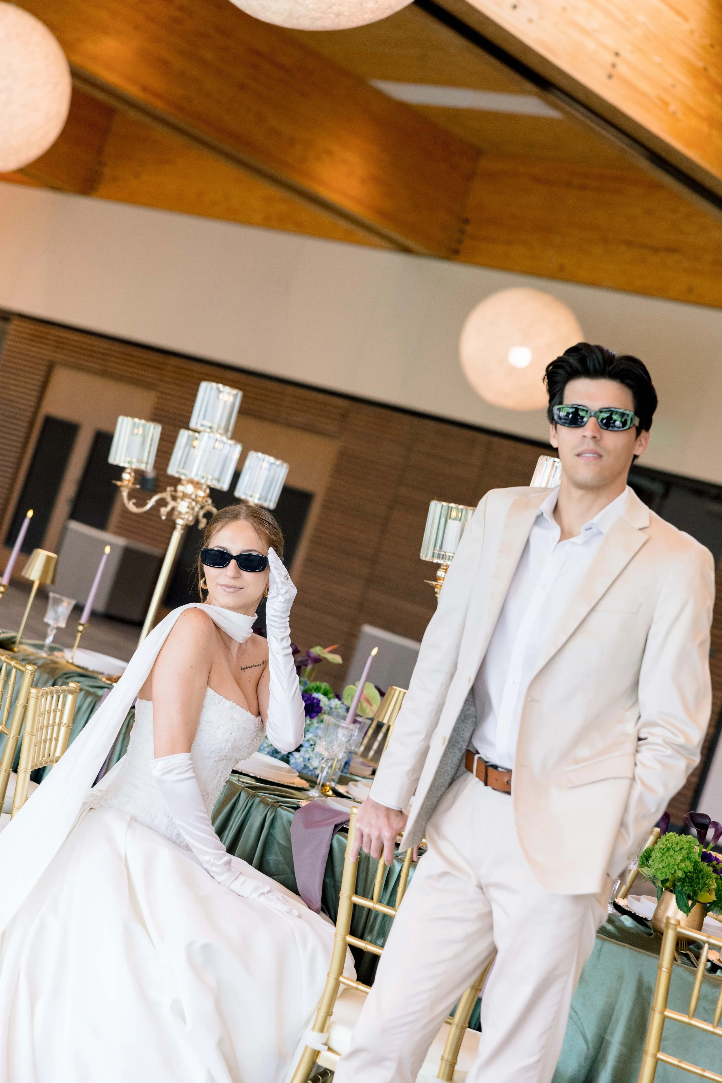 A woman in a white wedding dress and gloves, wearing sunglasses, seated at a table, with a woman standing next to a man in a cream-colored suit, also wearing sunglasses, at a reception with elegant table settings and golden chairs.