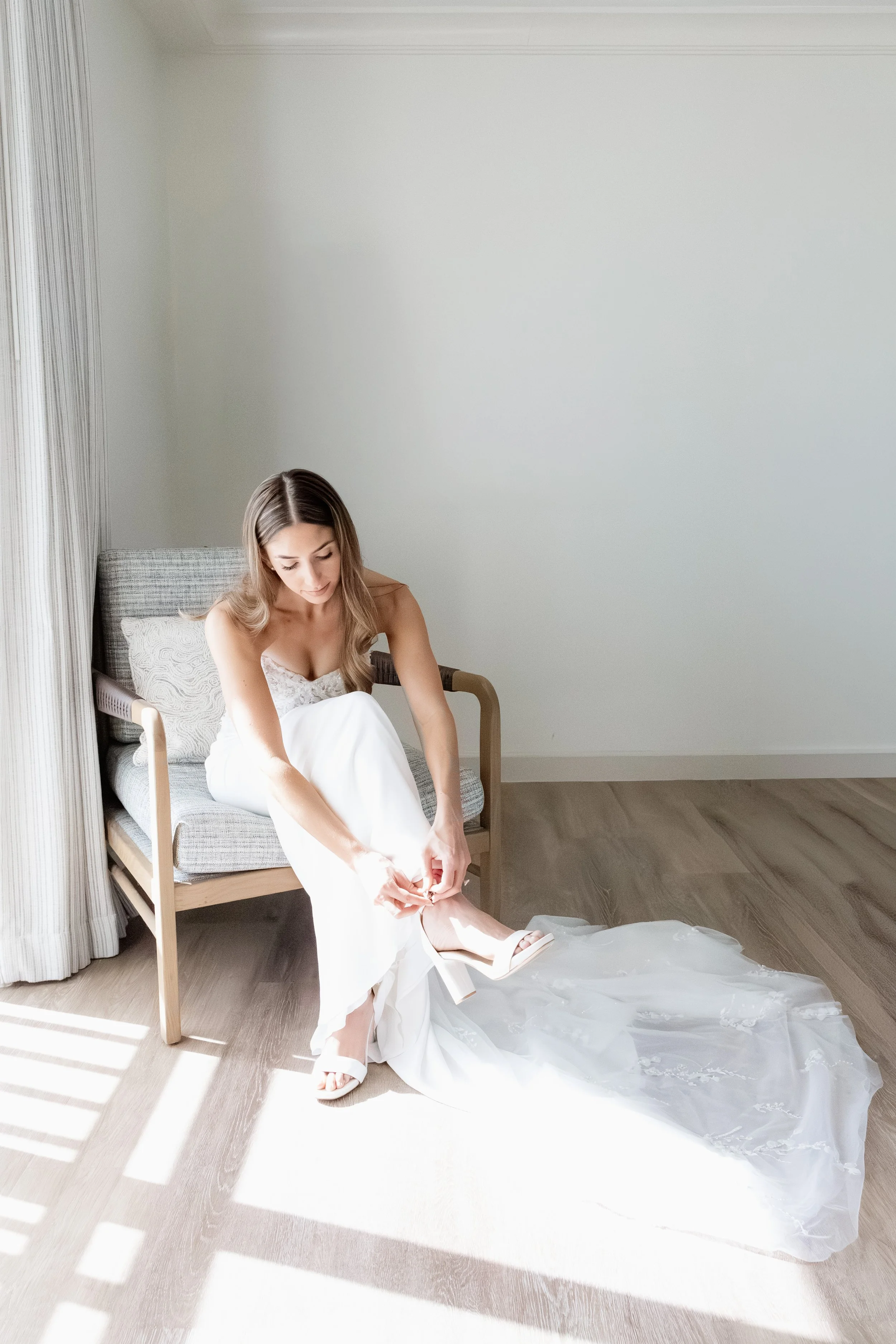 Bride in a wedding dress sits on a chair and puts on a white high heel shoe in a bright, modern room with wooden floors and abstract wall art at the Hyatt Regency in Bonita Springs, Florida 