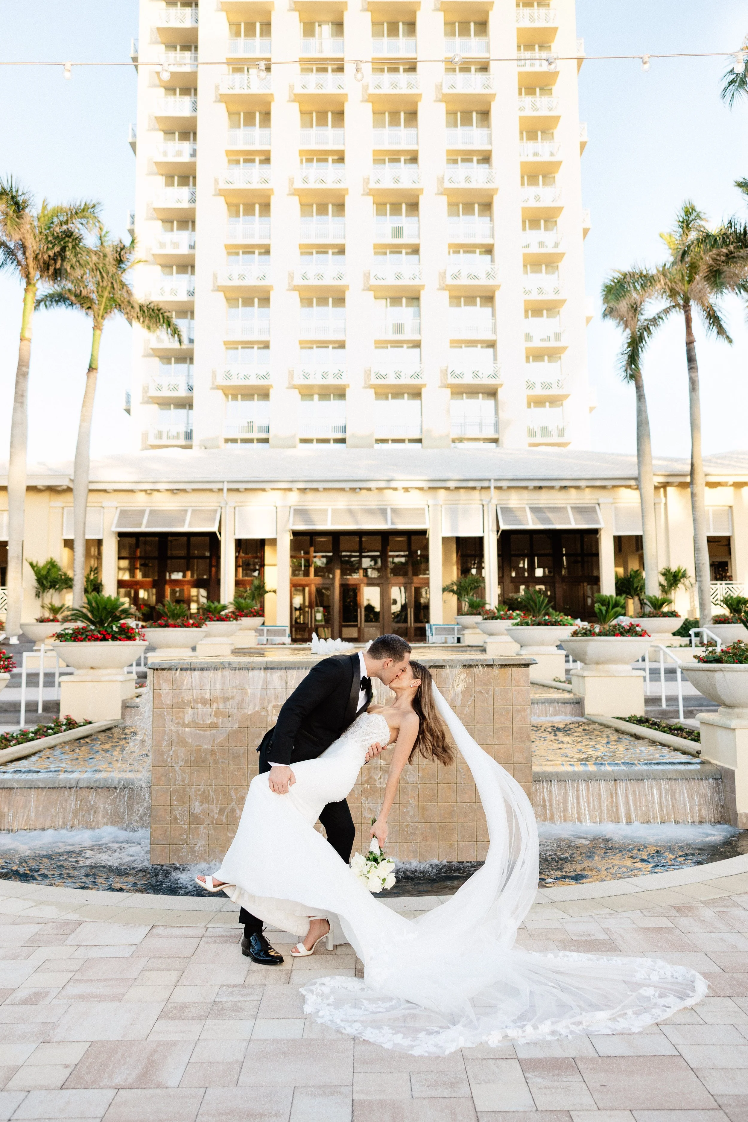 A newlywed couple sharing a kiss in front of a fountain at a hotel, the groom dips the bride as she holds a bouquet, and the bride wears a white wedding gown with a long veil, with a tall building, palm trees, and hotel entrance in the background 
