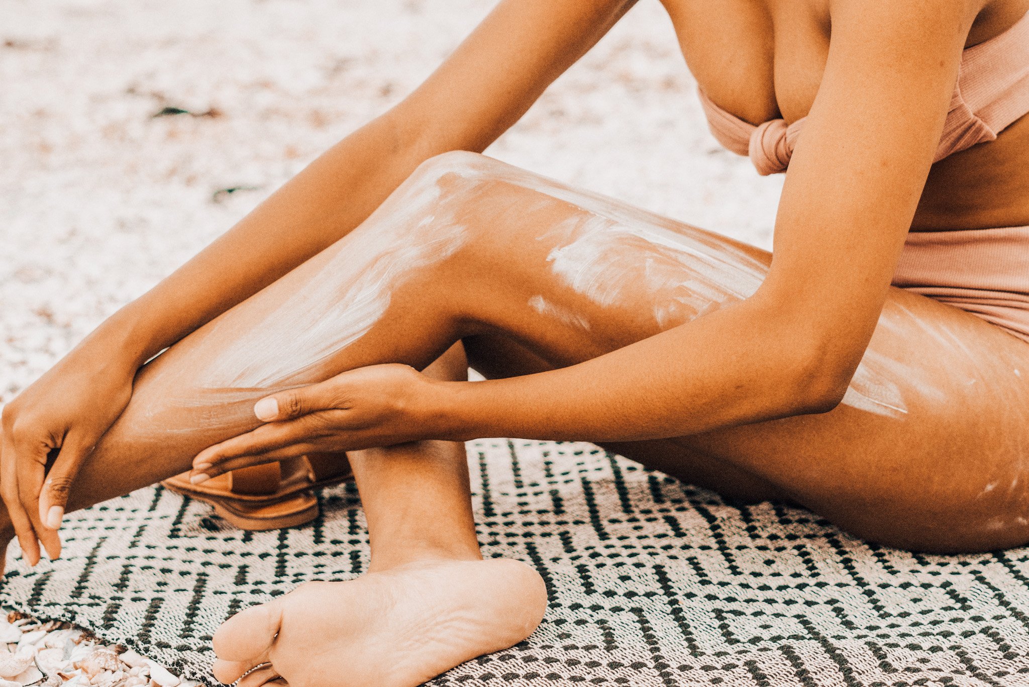 Person applying lotion to legs while sitting on a patterned beach mat, wearing a swimsuit.