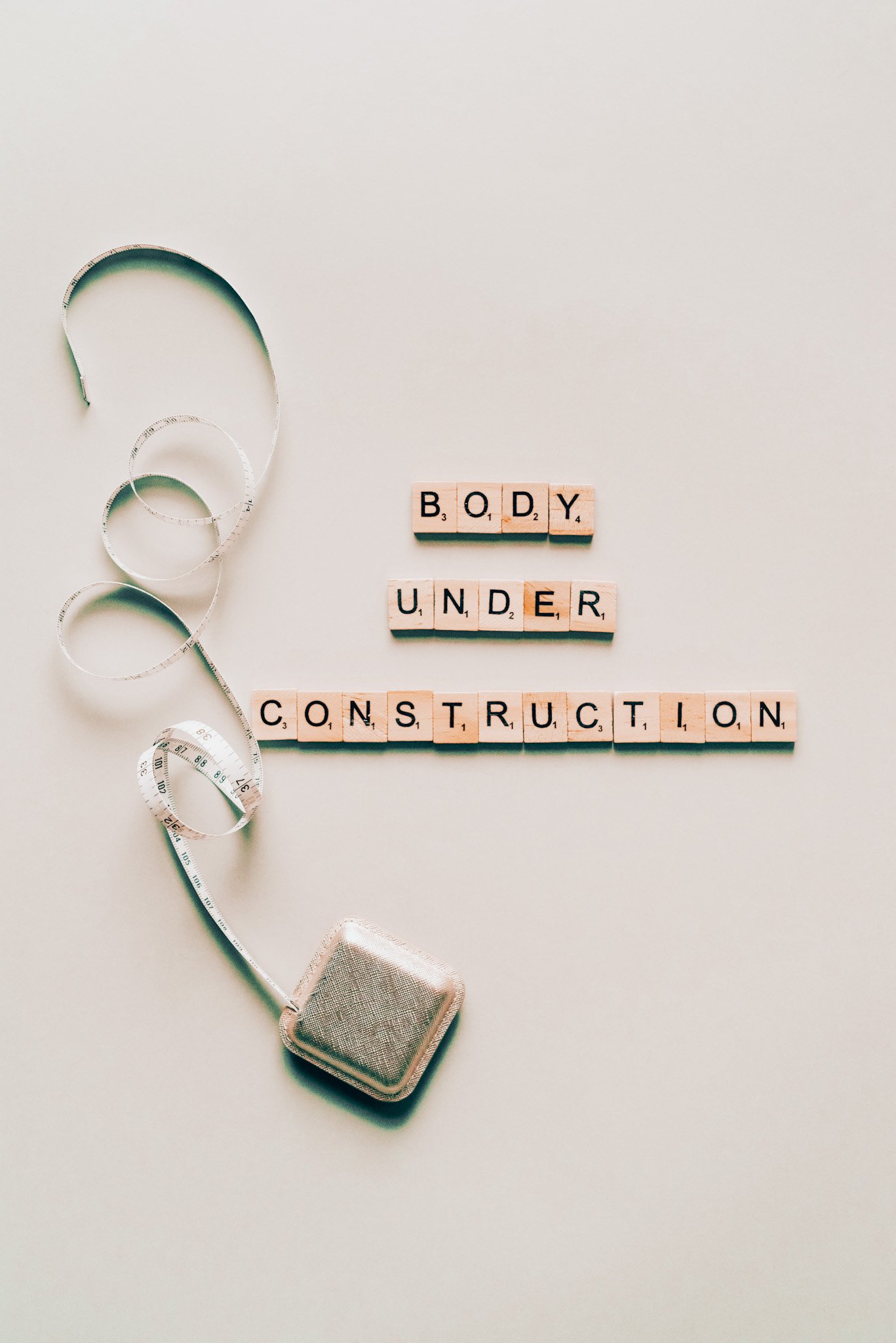 Scrabble tiles spelling 'BODY UNDER CONSTRUCTION' next to a coiled measuring tape on a beige background.