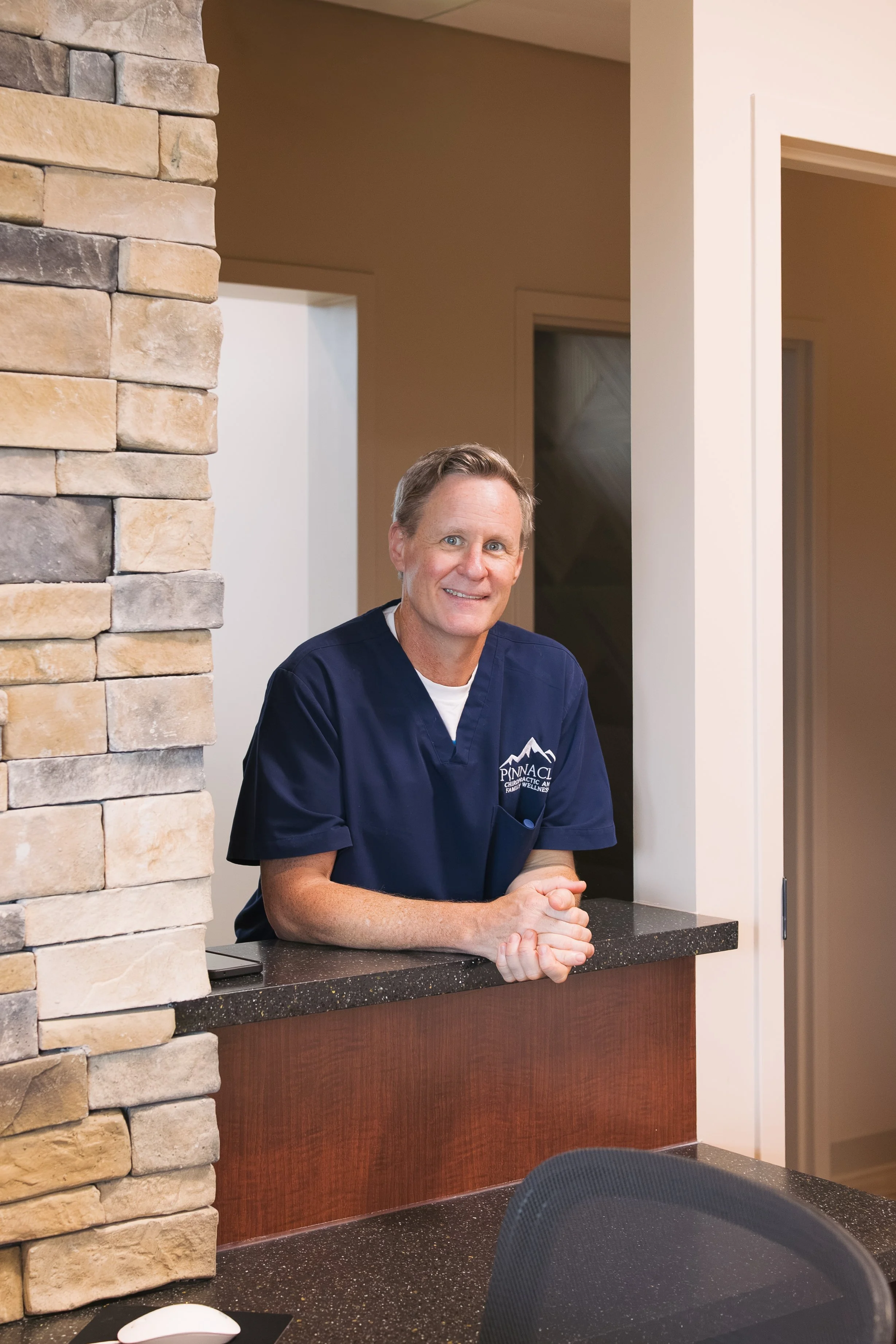 A man in medical scrubs standing indoors, smiling. The scrubs have a logo that reads Pinnacle Chiropractic and Family Wellness.