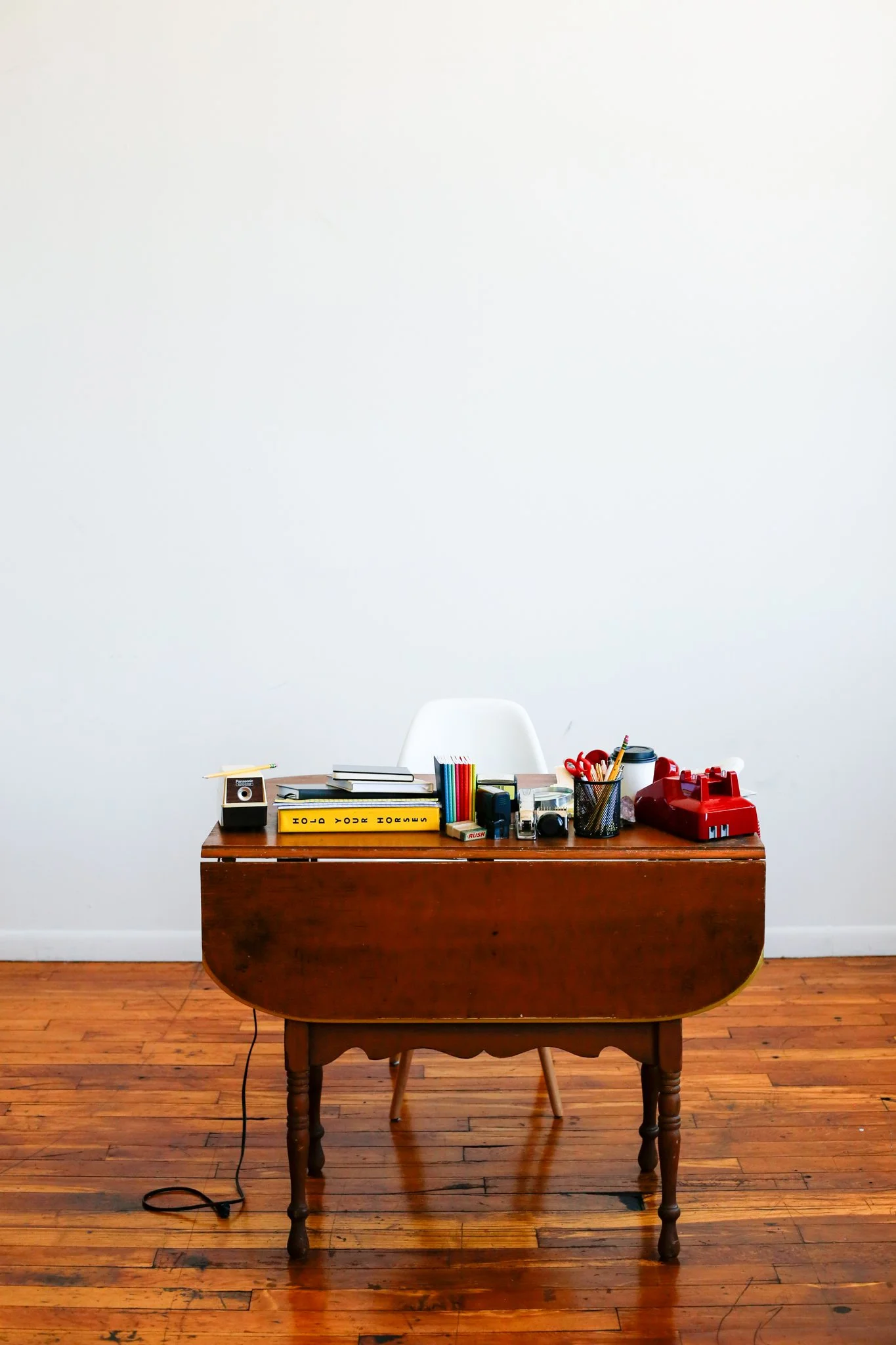Gosho Creative Photoshoot - Empty vintage wooden desk with various office supplies including books, colored pencils, scissors, camera, and a red typewriter, set against a plain white wall and hardwood floor.