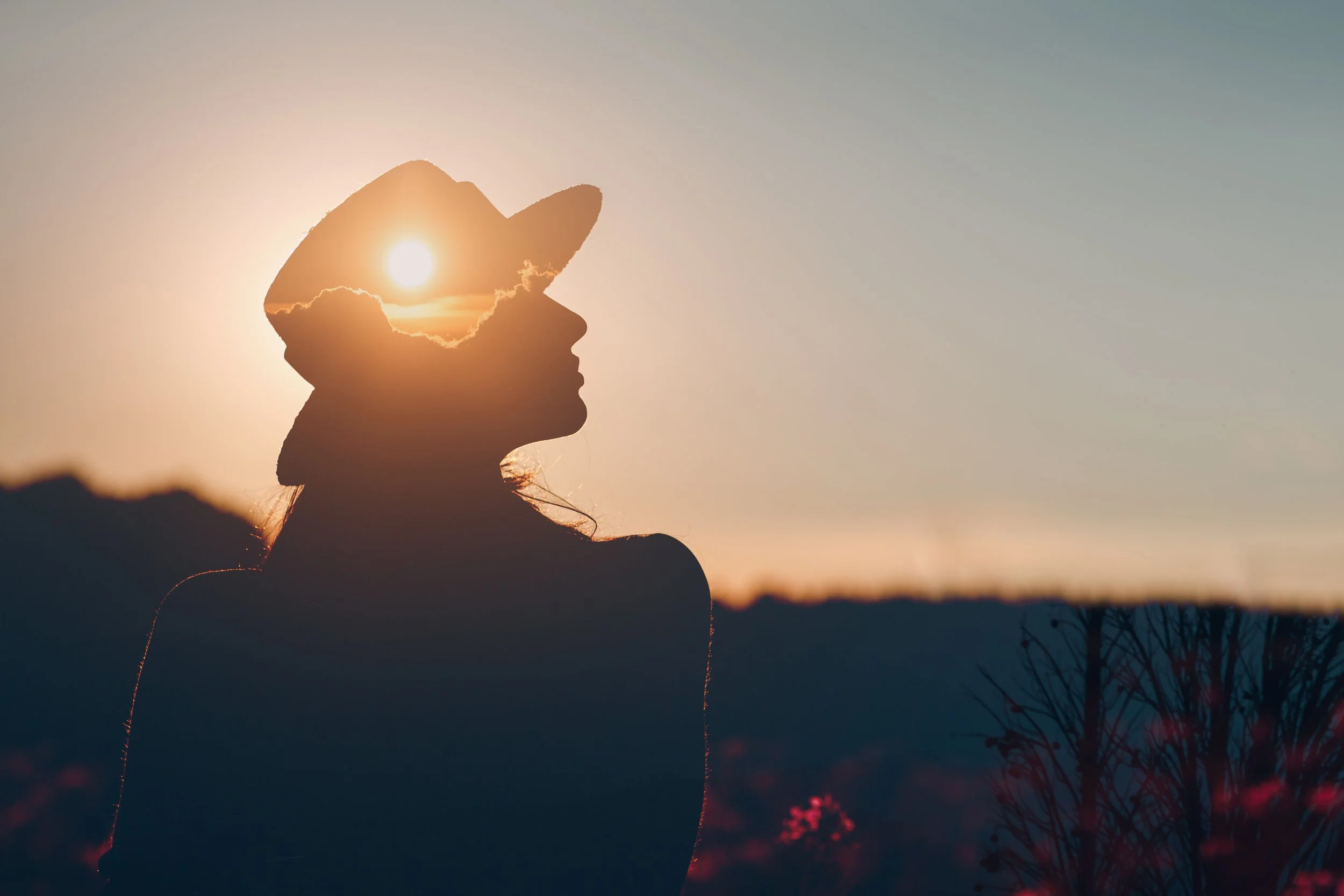 A silhouette of a woman wearing a hat, with the sun and clouds inside the hat’s outline during sunset, creating a double exposure effect.