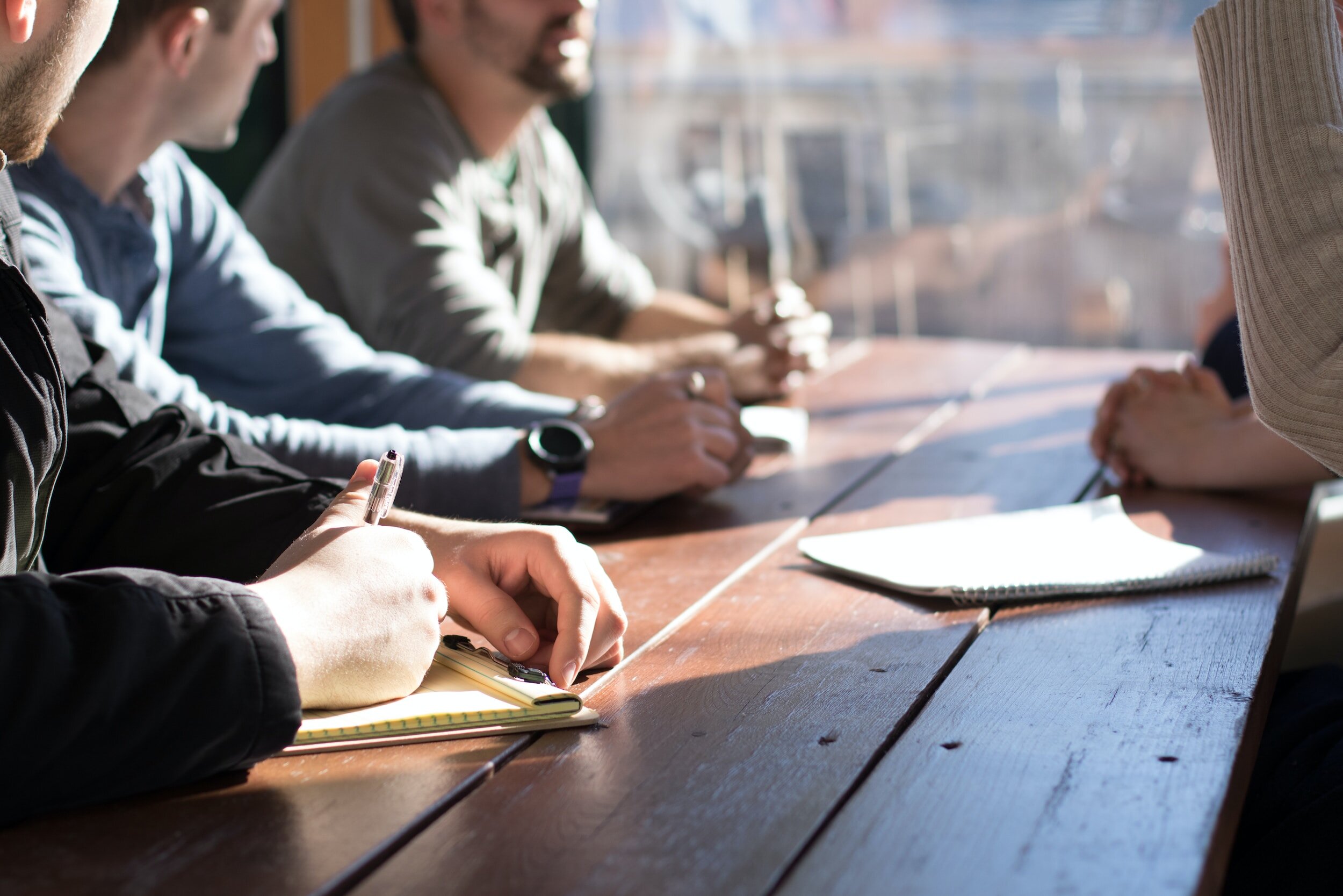 People sitting at a wooden table in a meeting or discussion, with notebooks and writing utensils in front of them, illuminated by natural light.